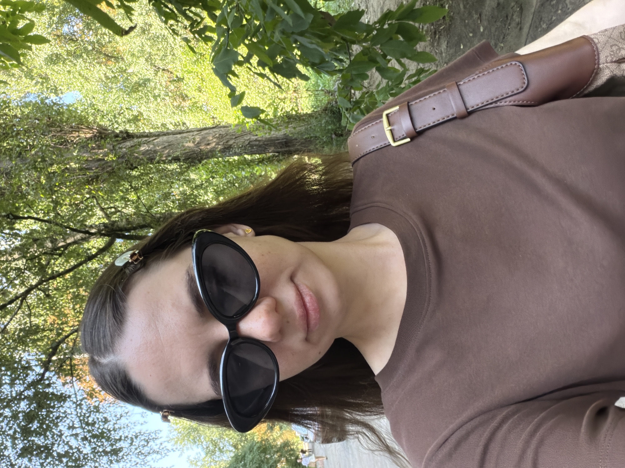 The image is a selfie of a young woman with dark hair, wearing black cat-eye sunglasses and a brown t-shirt. She is outdoors, with trees and foliage in the background, suggesting a park or wooded area within the city of Kyiv, Ukraine. A brown leather bag strap is visible over her right shoulder. In the background, two individuals are walking on a paved path, and a vehicle is partially visible. The lighting indicates it is daytime.
