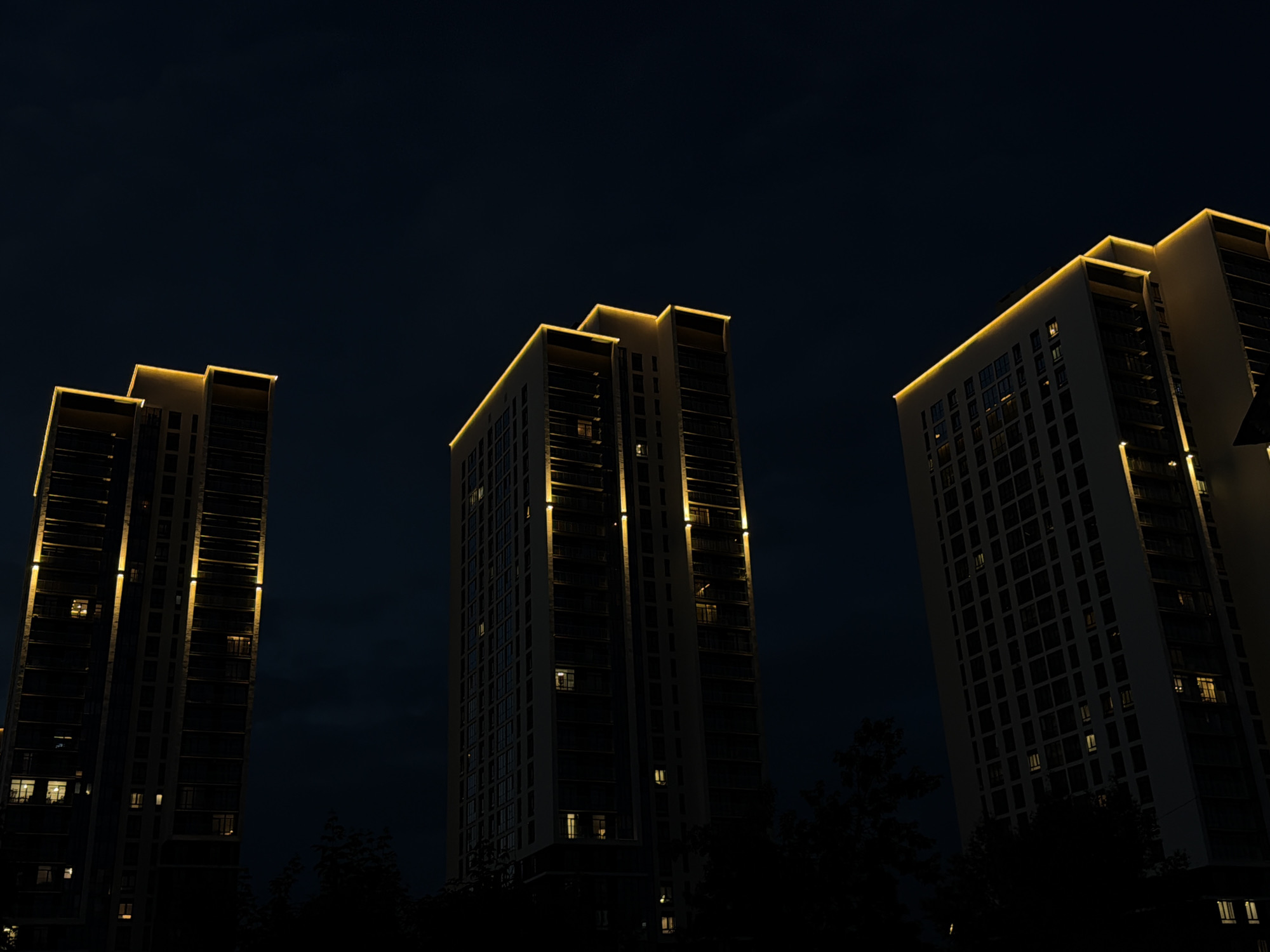 The image depicts three modern high-rise apartment buildings silhouetted against a dark night sky. Each building is illuminated with warm, golden lights along its edges and vertical accents, creating a striking contrast with the deep blue of the sky. The windows of the buildings are visible, with some showing interior lights, suggesting occupancy. The foreground is dark, with the lower portions of the buildings and some tree foliage obscured by shadow. The perspective is from below, looking up at the towering structures. The scene is characteristic of a contemporary urban landscape at night. This image was taken in Yekaterinburg, Russia.