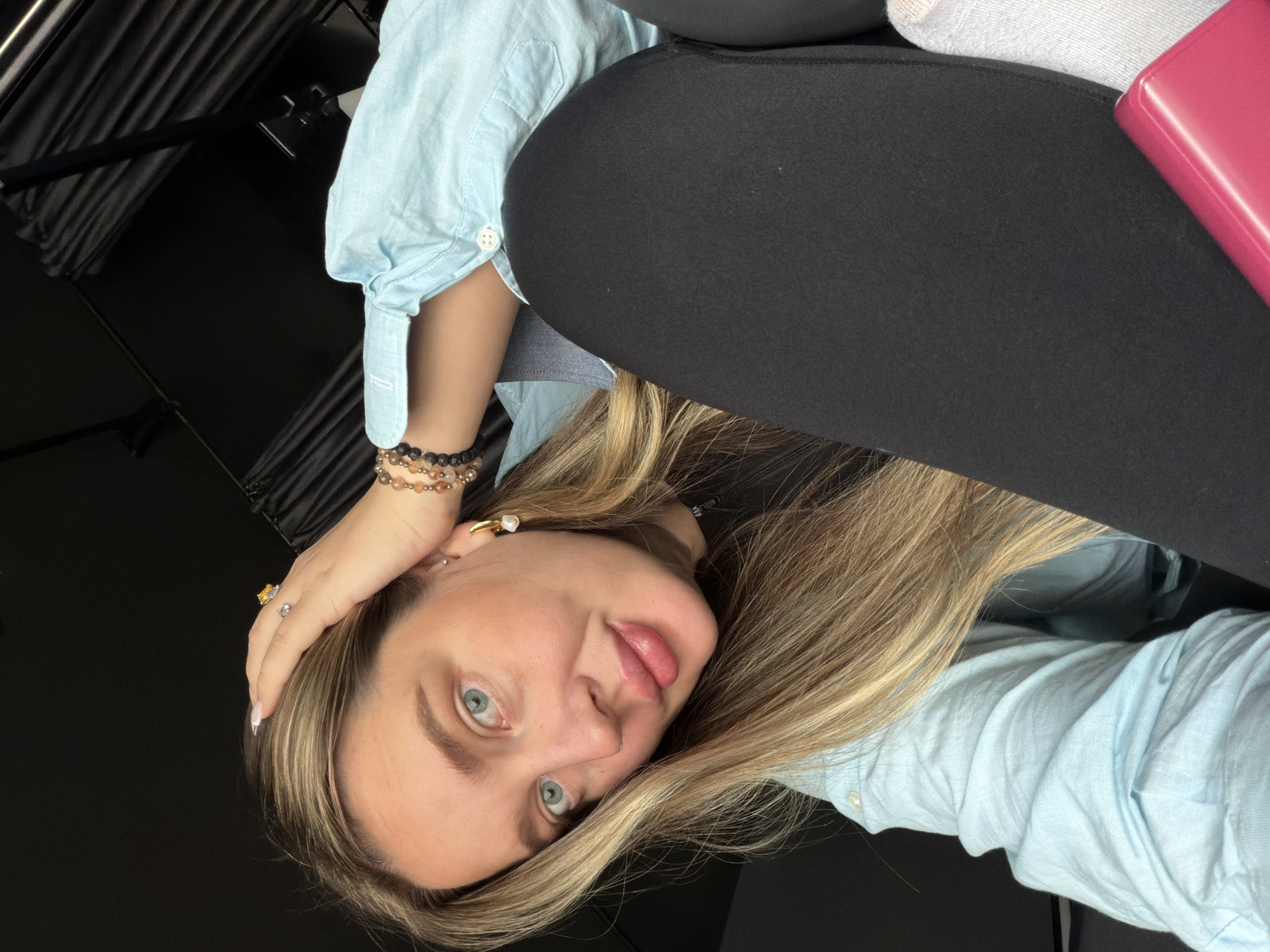A fair-skinned woman with blue eyes and blonde hair is looking at the camera. She is wearing a light blue button-down shirt and black pants. She has several rings on her left hand and bracelets on her left wrist. Her left hand is placed on her head. The background is dark, with a metallic lighting fixture on the left. There is also a pink object in the upper right corner of the image. The location in the city of Moscow, Russia, is not apparent from the image.