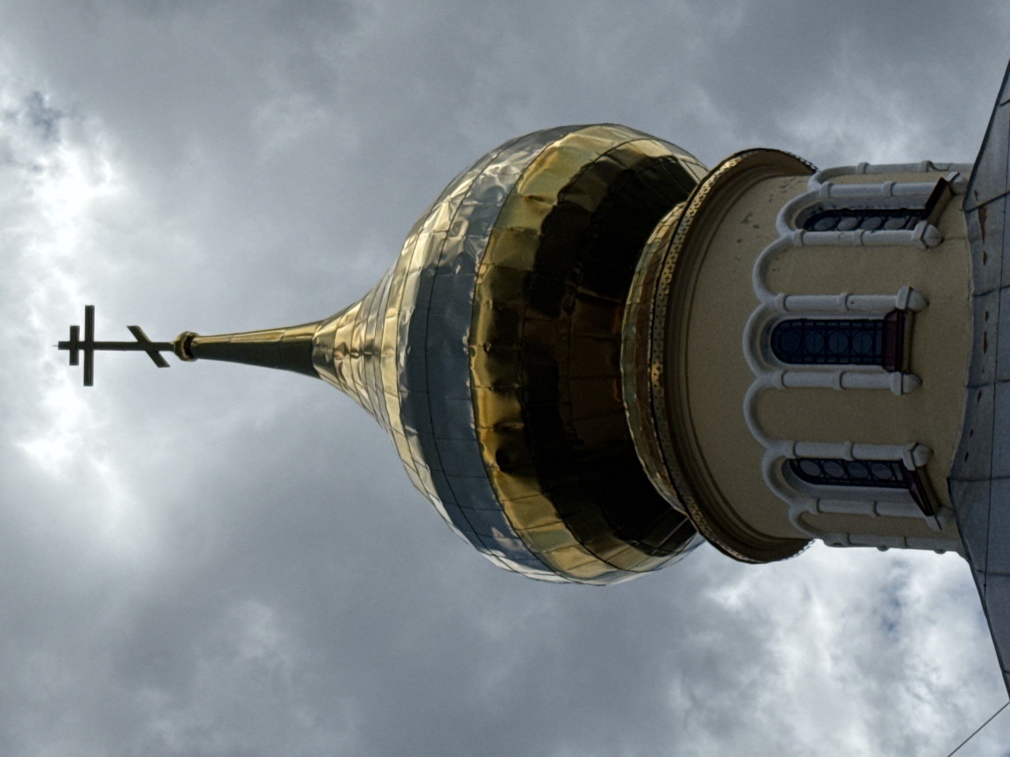 The image shows the golden dome and cross of an Orthodox church, silhouetted against a cloudy sky. The dome is bulbous and made of reflective material, with horizontal bands of darker and lighter tones. Below the dome is a cylindrical section of the church with arched windows. The cross at the apex of the dome is Eastern Orthodox, with a slanted lower bar. The overall impression is one of grandeur and religious significance. The specific location in Saint Petersburg, Russia, is not evident from the image alone.