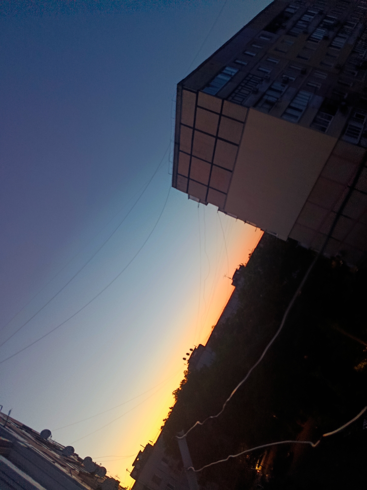 The image captures a tilted view of the sky and buildings at sunset or sunrise in Cherkaske, Ukraine. The sky transitions from a deep blue at the top to a soft orange and yellow near the horizon, indicating twilight. A multi-story building with numerous windows is prominently featured on the right side, its facade partially obscured by darkness. Below the building, trees and other structures are visible, silhouetted against the brightly lit sky. Several power lines stretch across the frame, adding a sense of urban infrastructure. On the left side of the image, another building or roofline with multiple satellite dishes is partially visible. The overall scene suggests an urban environment experiencing the transition from day to night or vice versa.