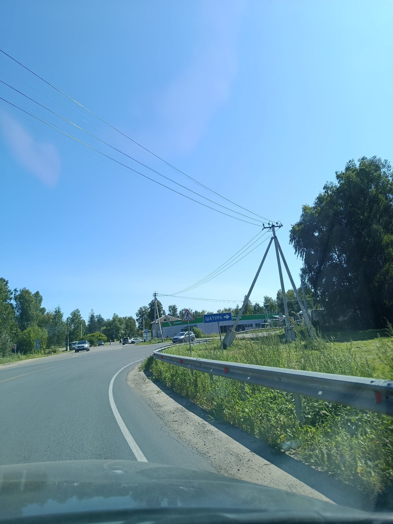 A road scene in Снопок Новый, Russia, features a winding asphalt road with a white lane marking. On the right side of the road, a metal guardrail runs alongside a grassy embankment. Power lines are strung between utility poles, with one A-frame pole prominently visible. A green building with the word "ШАТУРА" (Shatura) and an arrow pointing right is situated near the road. Several vehicles, including cars and what appears to be a truck or van, are present on the road and in the vicinity of the building. Trees and foliage line the sides of the road, and the sky is clear blue. The photograph is taken from the perspective of someone inside a vehicle, looking forward and slightly to the right.