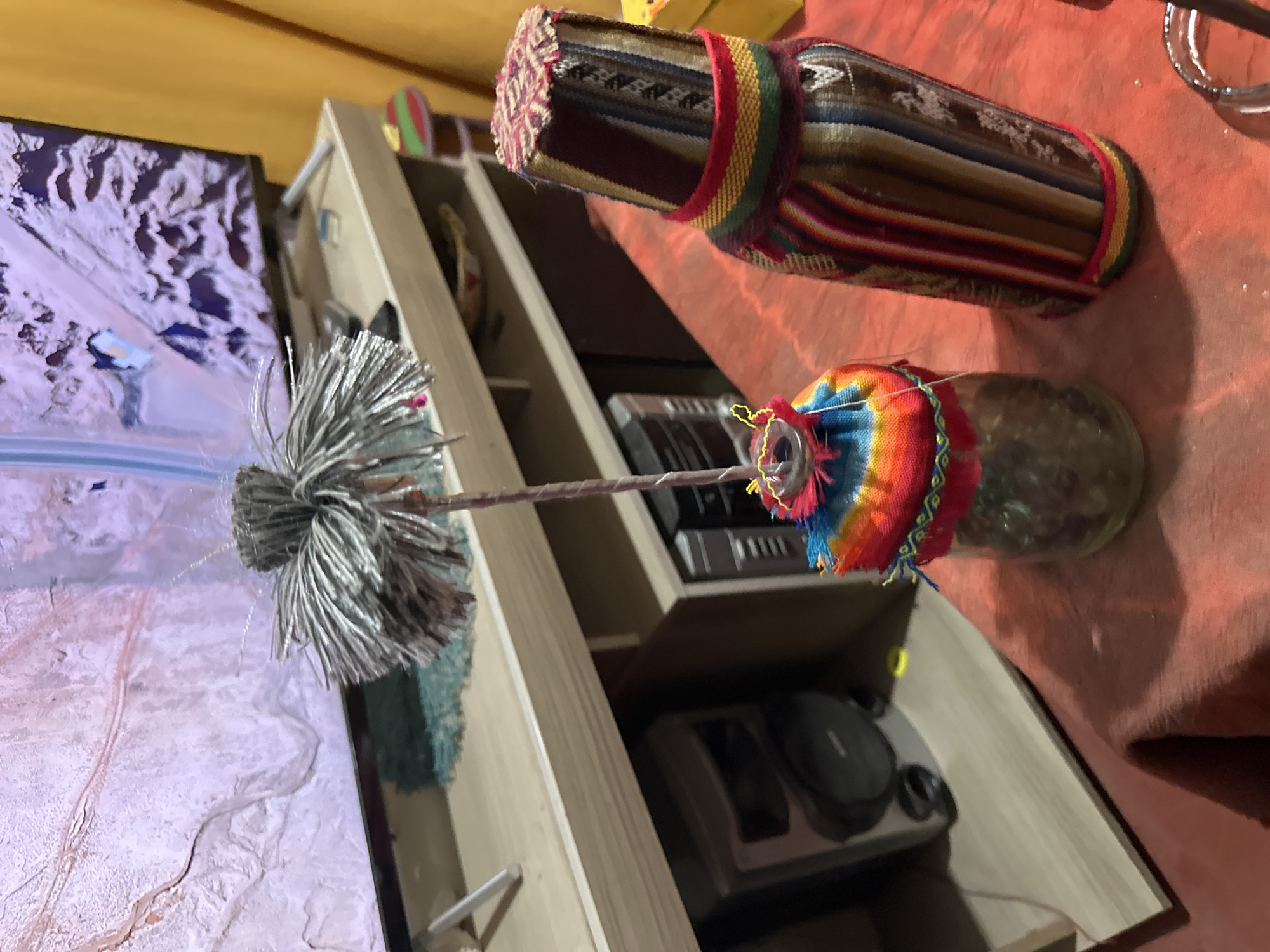 The image shows a living room setting with a television on the left displaying what appears to be a topographical map or satellite imagery. To the right of the television is a light-colored wooden shelving unit. On the shelves are various decorative items, including a bottle wrapped in colorful fabric, a decorative whisk-like object with silver strands, and what seem to be electronic devices, possibly media players or game consoles. The background features an orange-colored surface, likely a tablecloth or draped fabric.
Without additional context, it is impossible to determine if the image was taken in Arica, Chile. The objects in the scene, such as the fabric-wrapped bottle and the decorative whisk, could be indicative of Andean crafts, which are commonly found in Arica, but they could also be from another location.