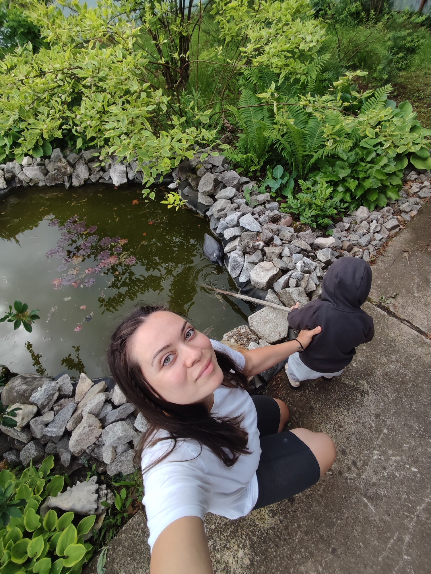 An adult female and a young child are situated outdoors beside a small, irregularly shaped pond in Фоки, Russia. The female, positioned on a concrete path in the foreground, is taking a selfie, looking directly at the camera. She wears a white t-shirt and dark shorts, with her left hand resting on the child's back. The child, wearing a dark hooded sweatshirt and light-colored pants, crouches on the concrete path, facing away from the camera towards the pond. The child holds a thin stick, which extends into the murky, dark green pond water. The pond is edged with numerous light gray rocks, and a dark pond liner is visible beneath them. The water's surface reflects an overcast sky and features reddish-purple water lily pads along with other green aquatic plants. The surrounding environment is lush with various green foliage, including a prominent variegated shrub with light green leaves, ferns, and hostas, with denser greenery in the background.