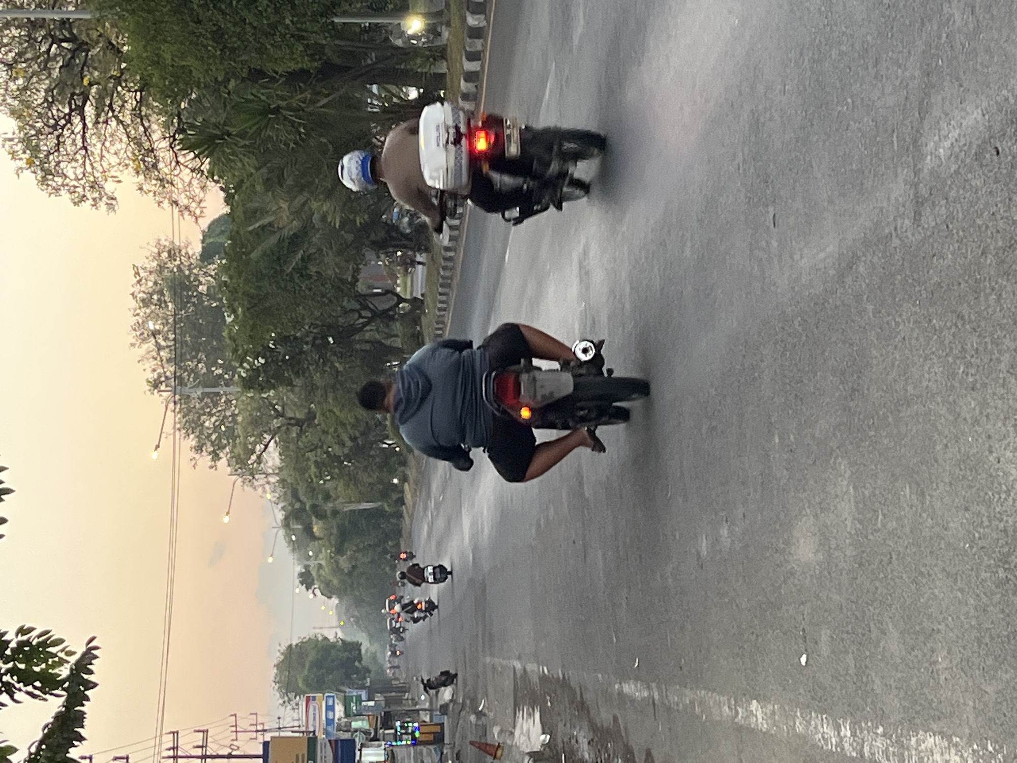 The image depicts a street scene in Ngaban, Indonesia, likely during sunset or early morning given the warm glow of the sky. Two men are riding motorcycles on a paved road. The man in the foreground, closest to the camera, is riding a motorcycle and is wearing a grey hooded sweatshirt and dark shorts. He is facing away from the camera. The second man is riding a motorcycle further down the road, also facing away from the camera. He is wearing a light-colored shirt and a blue and white helmet. Both motorcycles have their taillights illuminated. The road is lined with trees on both sides, and there are power lines and streetlights visible. On the left side of the road, there are buildings and some shops. The overall environment suggests a moderately busy street.