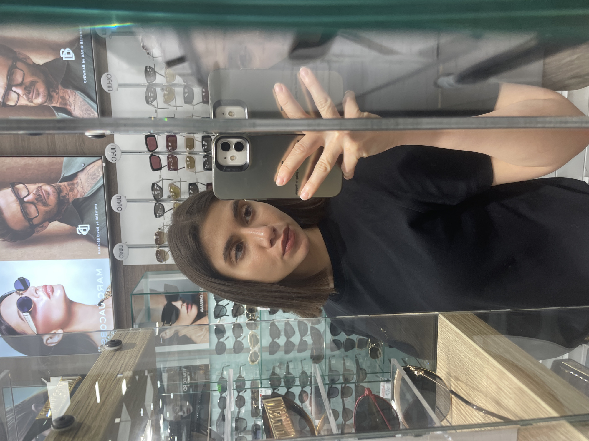 A woman is taking a selfie in front of a glass display case filled with sunglasses. The reflection shows rows of sunglasses and posters advertising brands like "Eyewear by David Beckham," "Liu Jo," and "Marc Jacobs." The location is likely an eyewear shop. There is no indication of location in Sofia, Bulgaria.