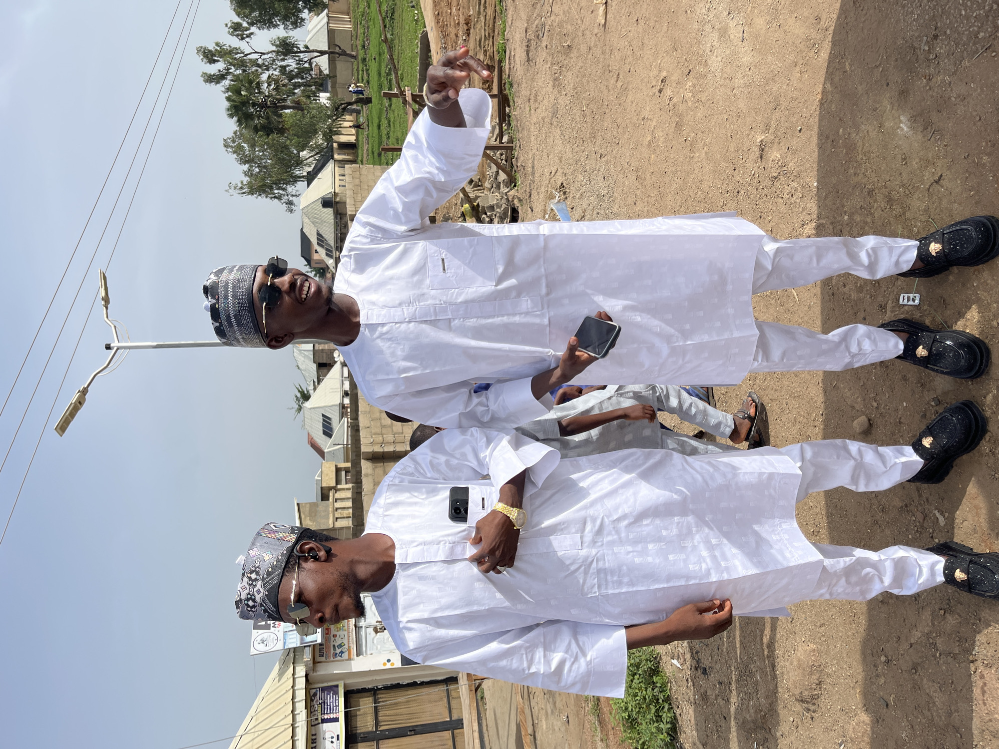 Two young men are posing for a picture outdoors on a dirt or gravel surface. They are both wearing similar traditional white outfits with matching hats and black shoes. One man is holding a phone, and the other is gesturing with his hand. In the background, there are buildings, trees, and a light pole. The location appears to be a city environment, possibly Jos, Nigeria.