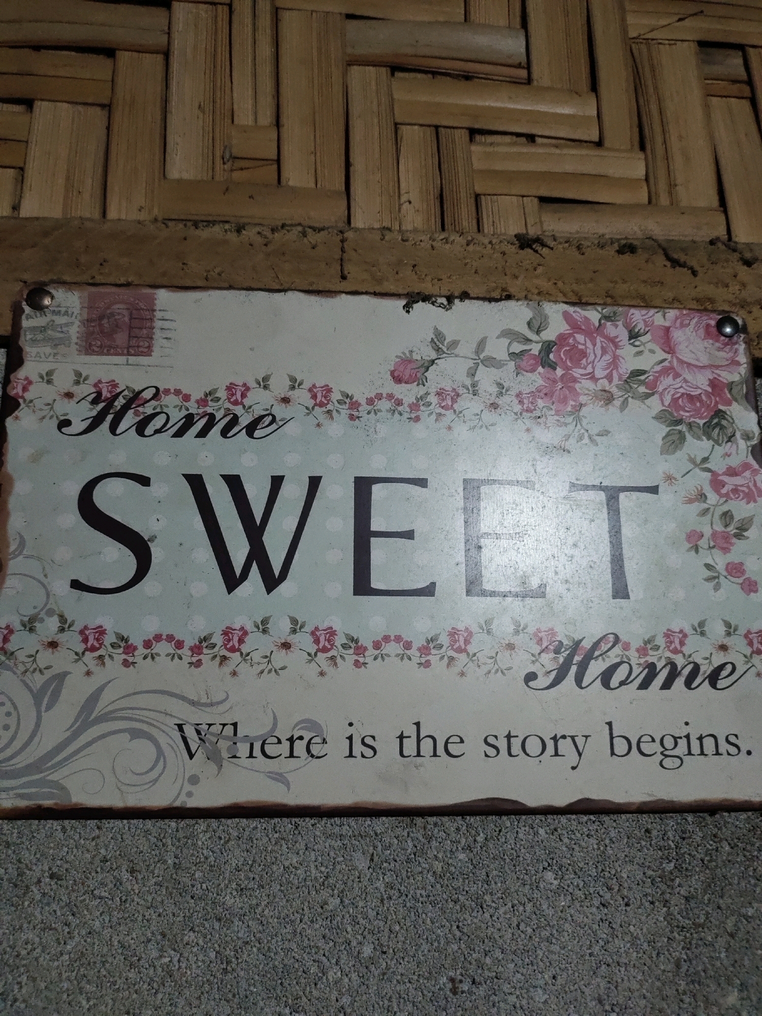 A distressed, rectangular sign with a floral pattern and the words "Home Sweet Home, Where is the story begins." is affixed to a woven bamboo wall. The sign has a light, aged finish with visible signs of wear and tear. The text is in a dark, contrasting font, with "Home Sweet" prominently displayed in large, bold letters. A smaller "Home" appears near the bottom right corner. The background of the sign features a light blue polka dot pattern and a border of pink roses and green foliage. The sign is secured with two visible metal brads on the top edge. The woven bamboo wall behind the sign is constructed of thin, light brown bamboo strips arranged in an interlocking pattern. The lighting suggests an indoor setting, possibly in a home or decorative display area within Matalam, Philippines. No people or animals are present in the image.