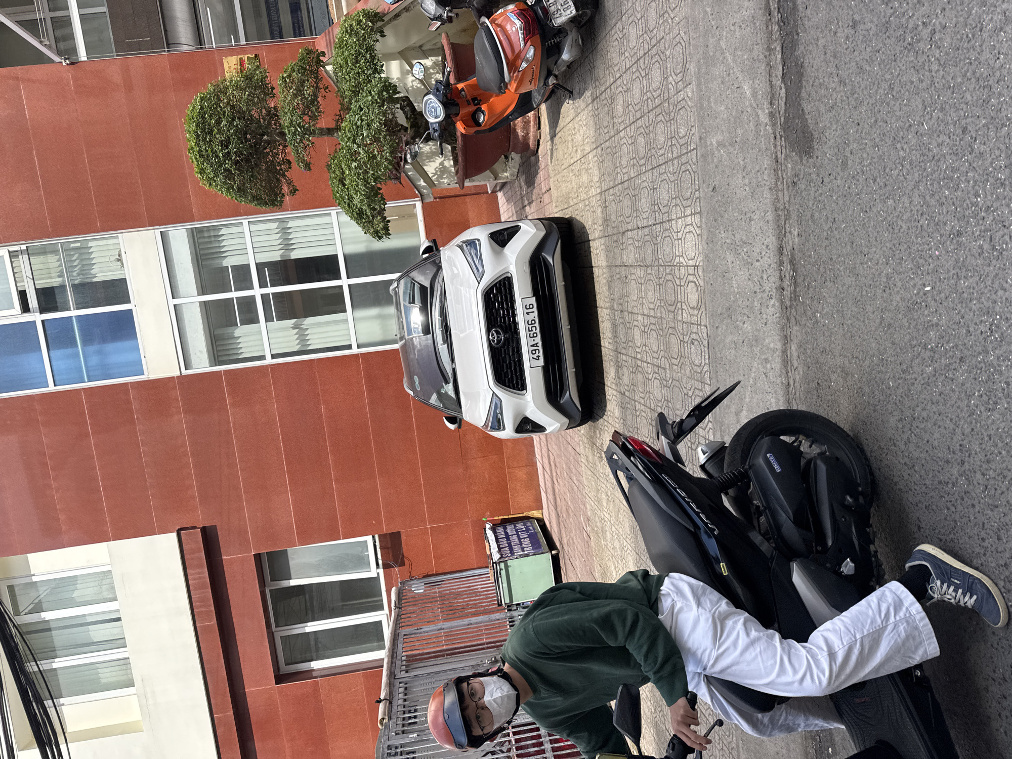 A man wearing a green sweater, white pants, a helmet, and a face mask rides a black scooter on a paved street in Da Lat, Vietnam. He is positioned in the foreground, facing left. Behind him, a white car with license plate "49A-656.16" is parked on a sidewalk. An orange and black scooter is also parked nearby. The background features a red building with windows and a decorative tree. The scene appears to be a sunny day.