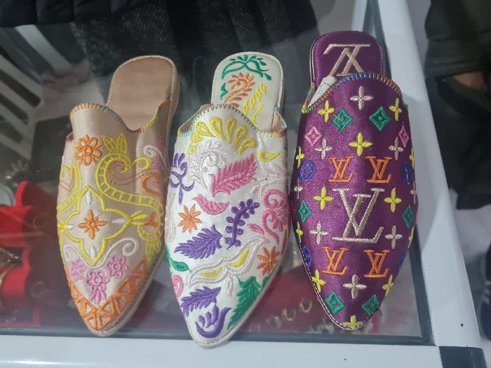 Three embroidered slippers are displayed. The left slipper features a light beige base with colorful floral and geometric patterns in orange, yellow, and pink. The middle slipper is white with an array of embroidered leaves and flowers in purple, pink, yellow, and green. The right slipper is purple and is decorated with an embroidered pattern of the Louis Vuitton logo in brown. They are arranged close together on a clear surface.