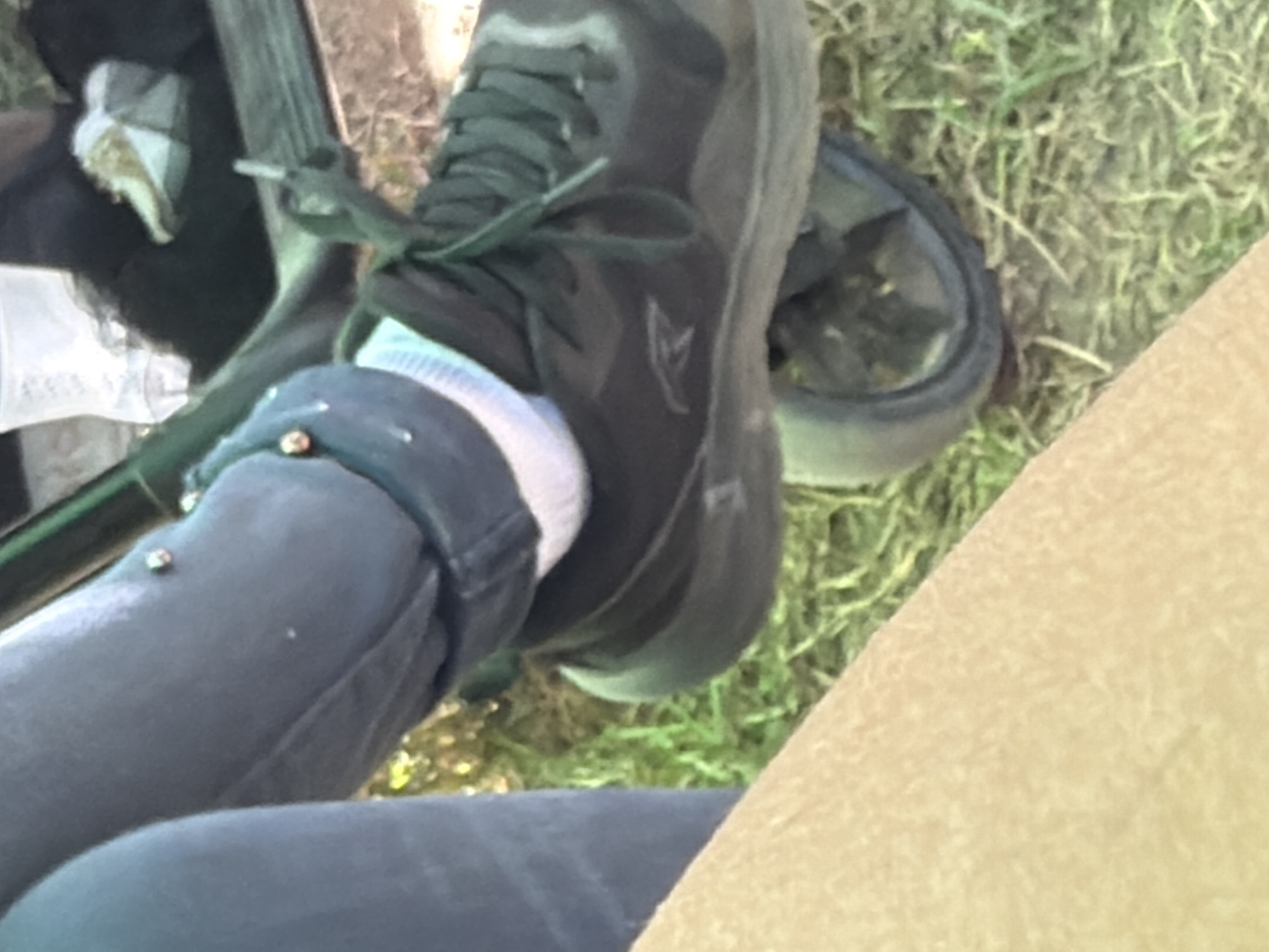 The image, taken in Razhevo Konare, Bulgaria, is a close-up, low-angle shot focused on a person's right leg and foot. The person is wearing dark blue pants that are embellished with small, reflective studs. A white sock with a light blue band is visible above a dark grey athletic shoe. The shoe features dark grey laces and a subtle logo on the side. The shoe is resting on a dark-colored wheel, which appears to be part of a stroller or similar wheeled object. The background consists of green grass and some dry leaves, suggesting an outdoor setting. The overall lighting is natural and appears to be daytime.
