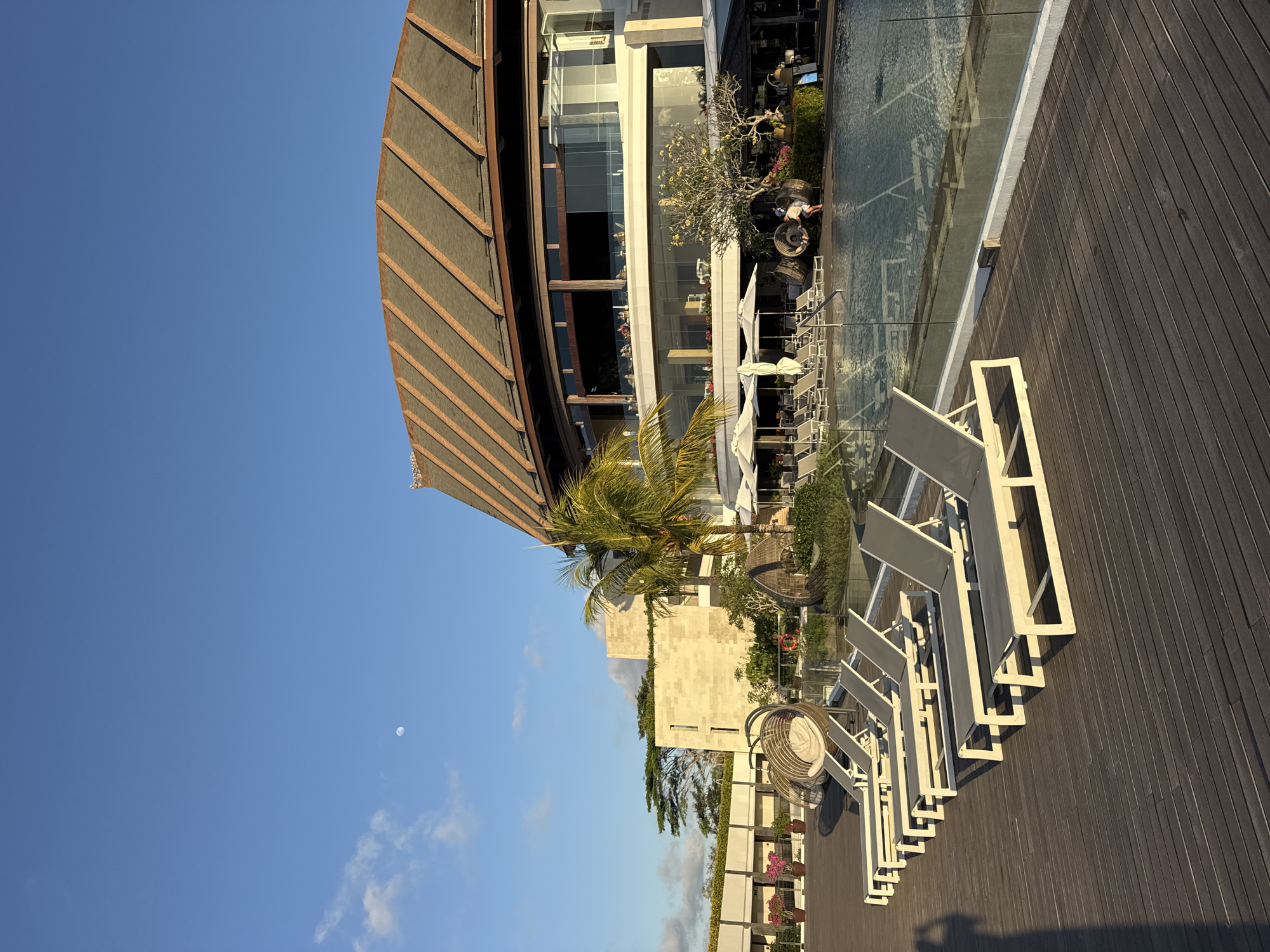 The image depicts a resort pool area in Kuta Selatan, Indonesia, under a clear blue sky with a visible moon. In the foreground, a wooden deck is lined with several empty lounge chairs. Beyond the chairs, a swimming pool is visible, bordered by a glass fence. To the right of the pool, a large, modern building with a distinctive curved, tiled roof stands prominently. The building features multiple levels with large windows. Palm trees and other tropical vegetation are scattered throughout the scene, suggesting a warm climate. A few people are present near the pool and in the building, but they are not the primary focus. The lighting suggests it is either early morning or late afternoon.