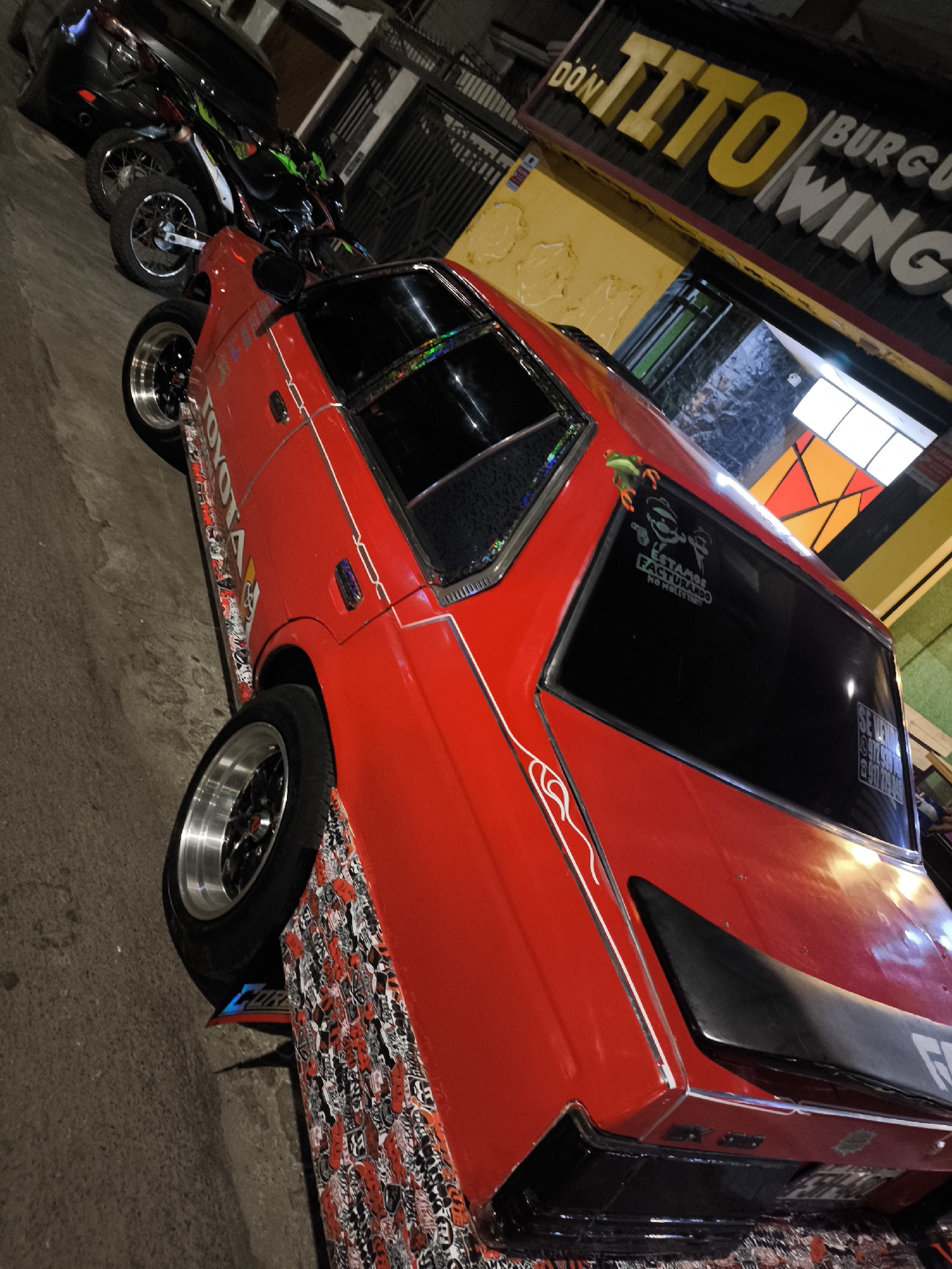 The image captures a red Toyota car parked on a city street at night. The car has various stickers, including a "TOYOTA" decal and another with a frog and cartoon figure, with the text "ESTAMOS FACTURANDO NO MOLESTAR!" on the rear window. There's also a "SE VENDE" sign on the window with a phone number. Below the door, a sticker bomb pattern covers the side.
Behind the red car, a motorcycle and a black car are also parked along the street. In the background, there is a building with a sign that reads "DON TITO BURGU/WINGS," suggesting a food establishment. The location appears to be an urban area at night.