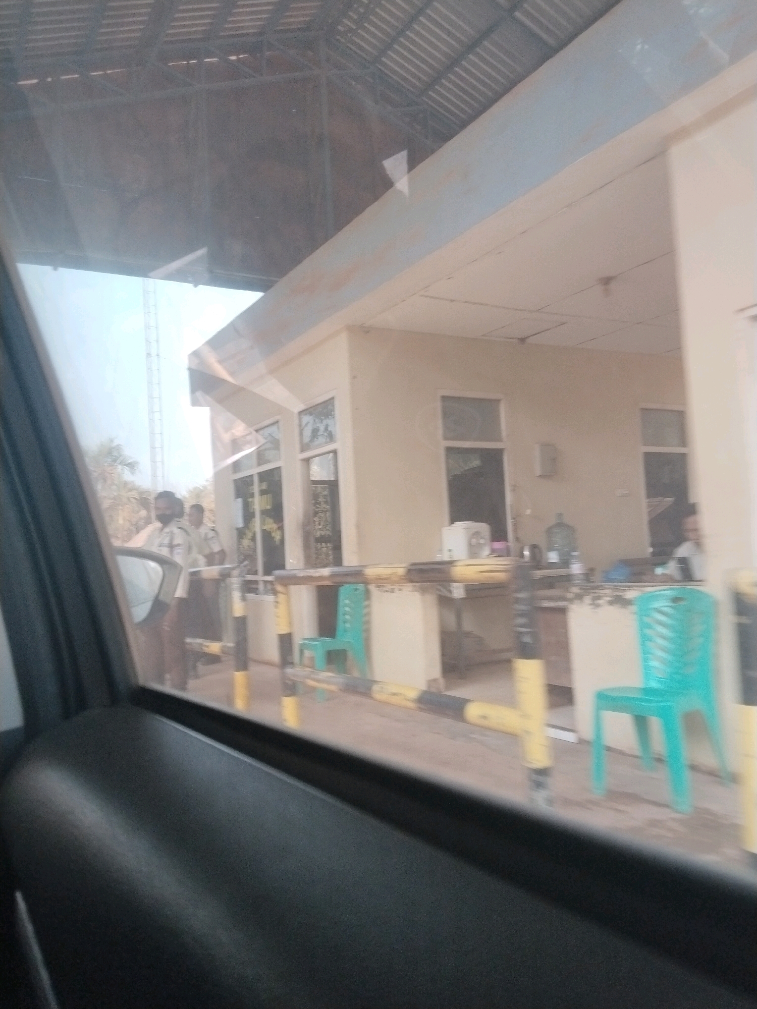 The image depicts an outdoor scene near a building with an overhang, likely a security checkpoint or an office, located in Dente Makmur, Indonesia. The perspective is from inside a vehicle, looking out through the side window. In the foreground, the dark interior of a car door is visible. Beyond the car window, there is a pathway with yellow and black striped bollards and railings. Two individuals, appearing to be security personnel wearing light-colored uniforms, are standing near the entrance of the building. The building itself is light-colored with several windows. In front of the building, there are two bright turquoise chairs. The environment suggests a public or semi-public space, possibly a checkpoint or an entrance to a facility.