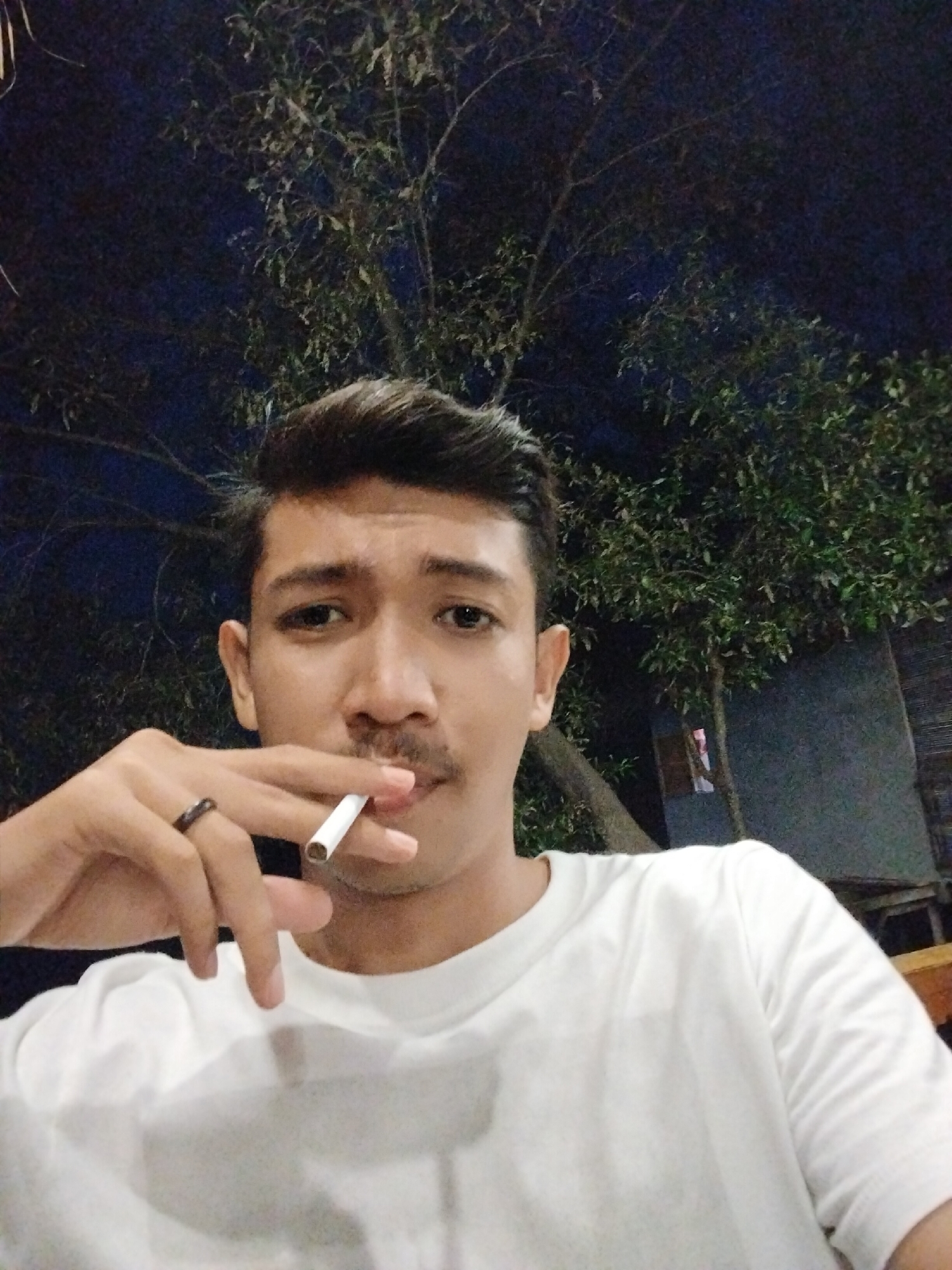 A young man with dark hair and a mustache is smoking a cigarette, holding it between his index and middle fingers. He is wearing a white t-shirt and a dark ring on his ring finger. The background is dark, with the silhouette of a tree and some foliage visible. The image appears to be taken at night, possibly in an outdoor setting. Based on the available information, the scene could be located in the city of Lhokseumawe, Indonesia.