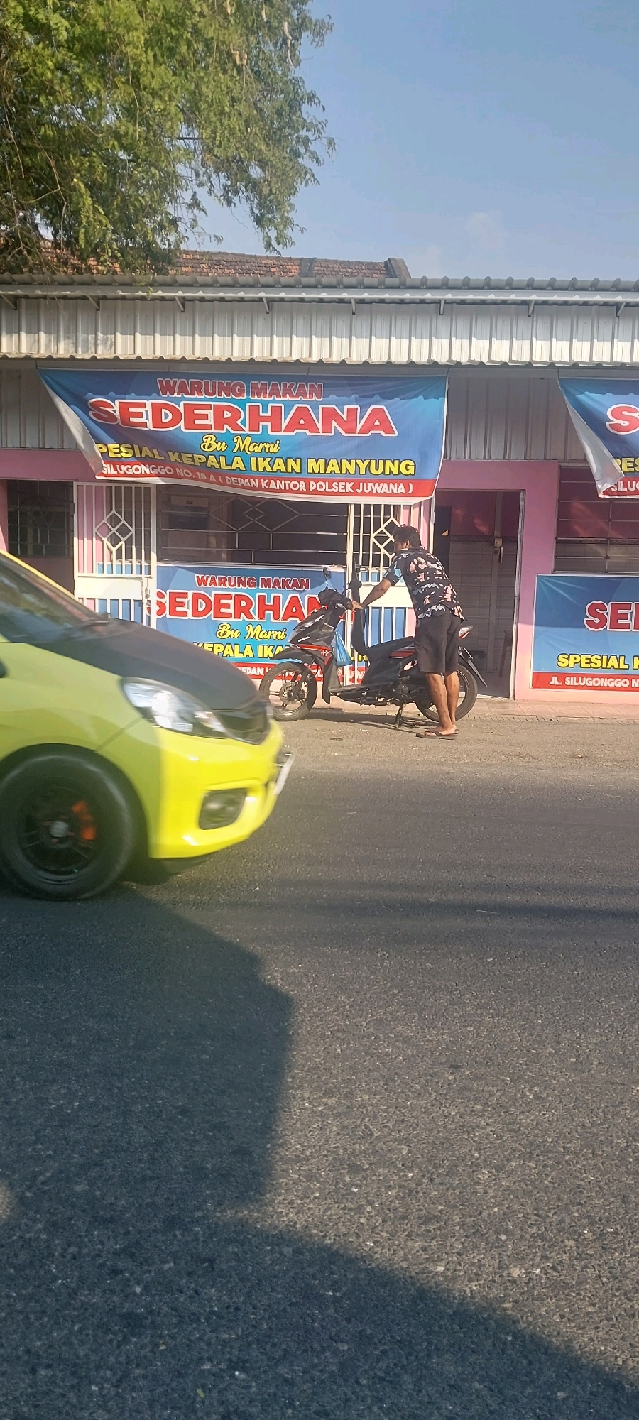 A bright yellow car is parked on the left side of the road, with its front bumper and headlight visible. The car is positioned at an angle, suggesting it might be moving or has recently stopped. In the background, a man wearing a dark patterned shirt and shorts is standing next to a black motorcycle, with his hand on the handlebars. He appears to be interacting with the motorcycle. Behind the man and motorcycle, there is a building with a pink facade and a white awning. Two blue banners are hung on the building's facade. The banner on the left reads "WARUNG MAKAN SEDERHANA" in large red letters, with smaller text below detailing "Spesial Kepala Ikan Manyung" and an address "Jl. Silugonggo No. 18 A (Depan Kantor Polsek Juwana)". A similar banner is partially visible on the right side of the building. The building's roof has a tiled structure, and a tree with green leaves is visible above the building on the left. The scene is illuminated by bright sunlight, casting shadows on the asphalt road. The location is identified as Juwana, Indonesia, with the building appearing to be a restaurant or food stall.