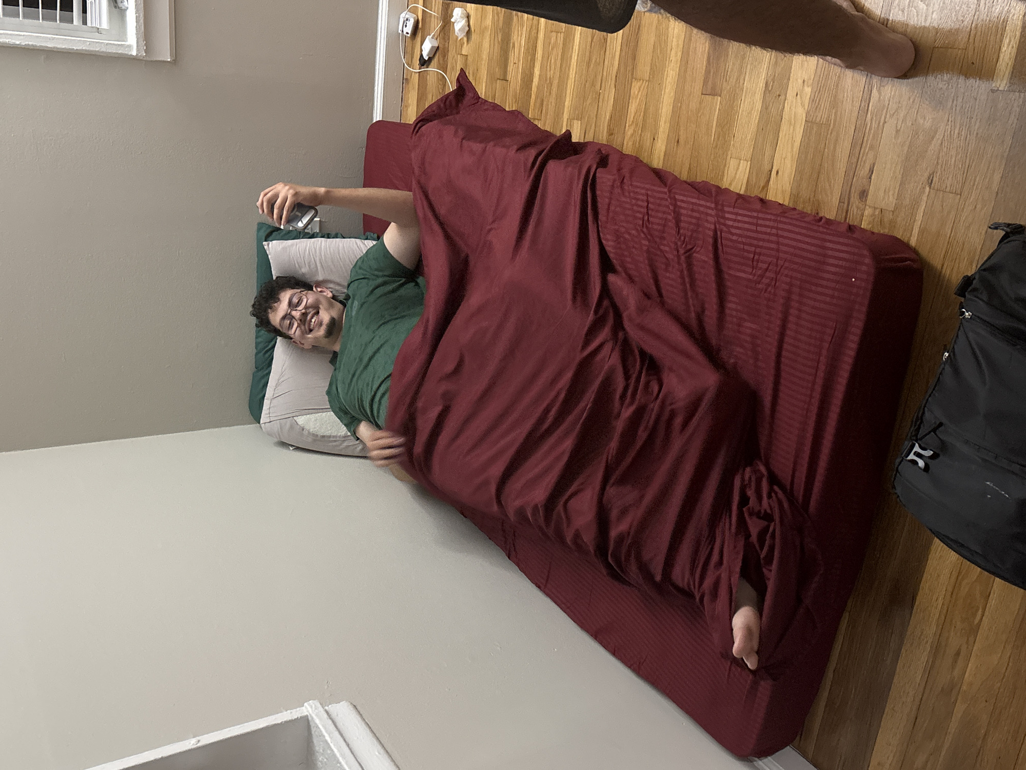 The image captures an indoor scene, likely a bedroom or temporary sleeping area, with a man lying on a bed. The main subject is a young man with dark, curly hair, a beard, and glasses. He is wearing a green t-shirt and is smiling broadly, appearing relaxed and happy. He is lying on a white pillow and is partially covered by a dark red duvet or blanket, which has subtle stripe patterns. He holds a silver-colored smartphone in his right hand, pointing it roughly towards himself, suggesting he might be taking a selfie or engaging in a video call. His bare left foot is visible, poking out from under the blanket at the foot of the bed.
The bed consists of a mattress covered with a dark red fitted sheet, placed directly on a light wooden floor. The room features light grey walls. A window with white blinds is visible in the upper left corner of the frame. In the upper right portion of the image, the bare legs of another person are visible, implying someone else is standing or walking nearby. On the wall near the head of the bed, a cluster of white power outlets with various cords plugged in is visible. A black backpack rests on the wooden floor beside the foot of the bed. The overall impression is candid and informal. No discernible text is visible in the image.