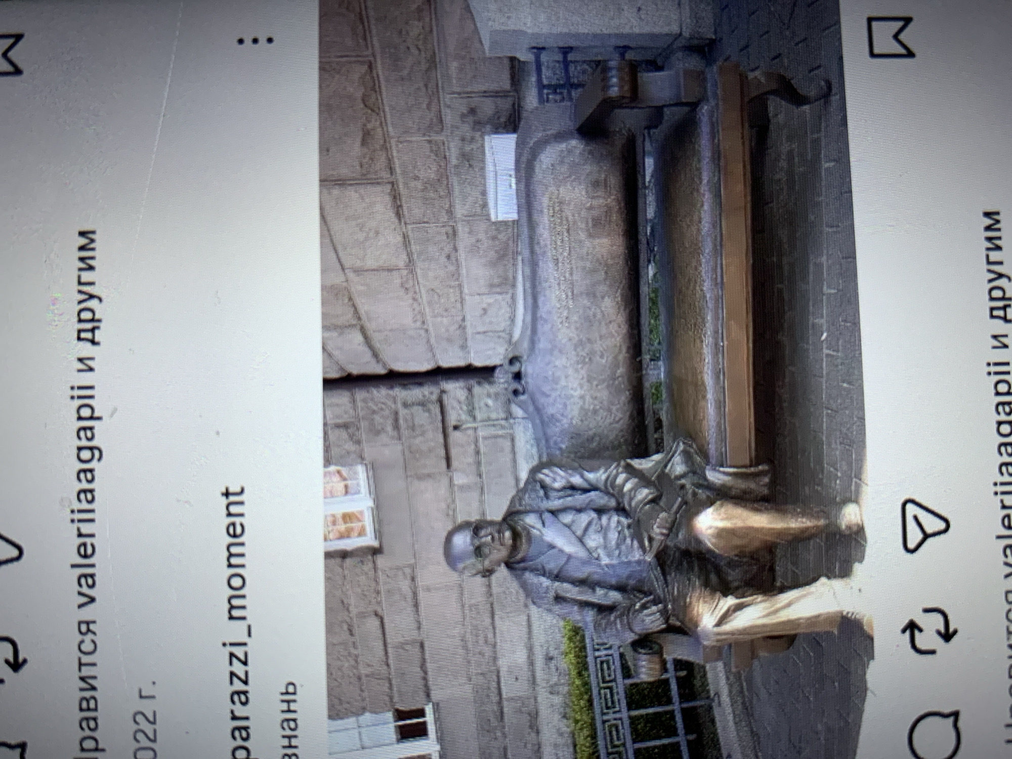 The image displays a statue of a man seated on a bench in an outdoor setting. The statue is made of a metallic material and depicts the man wearing a coat, shirt, and trousers, with glasses on his face. He is holding an object in his lap. The bench appears to be made of metal and wood. The background consists of a stone building and some greenery. The image is a photograph taken from a slightly elevated angle, possibly of a screen displaying an image. Text is visible at the top and bottom of the image, suggesting it is a screenshot from a social media platform. One line of text reads "нравится valeriiaagapii и другим" which translates to "likes valeriiaagapii and others". Another line reads "2022 г." indicating the year 2022. The text "parazzi_moment" is also visible. There are no elements in the image that definitively place the scene in the city of Lymanka, Ukraine.