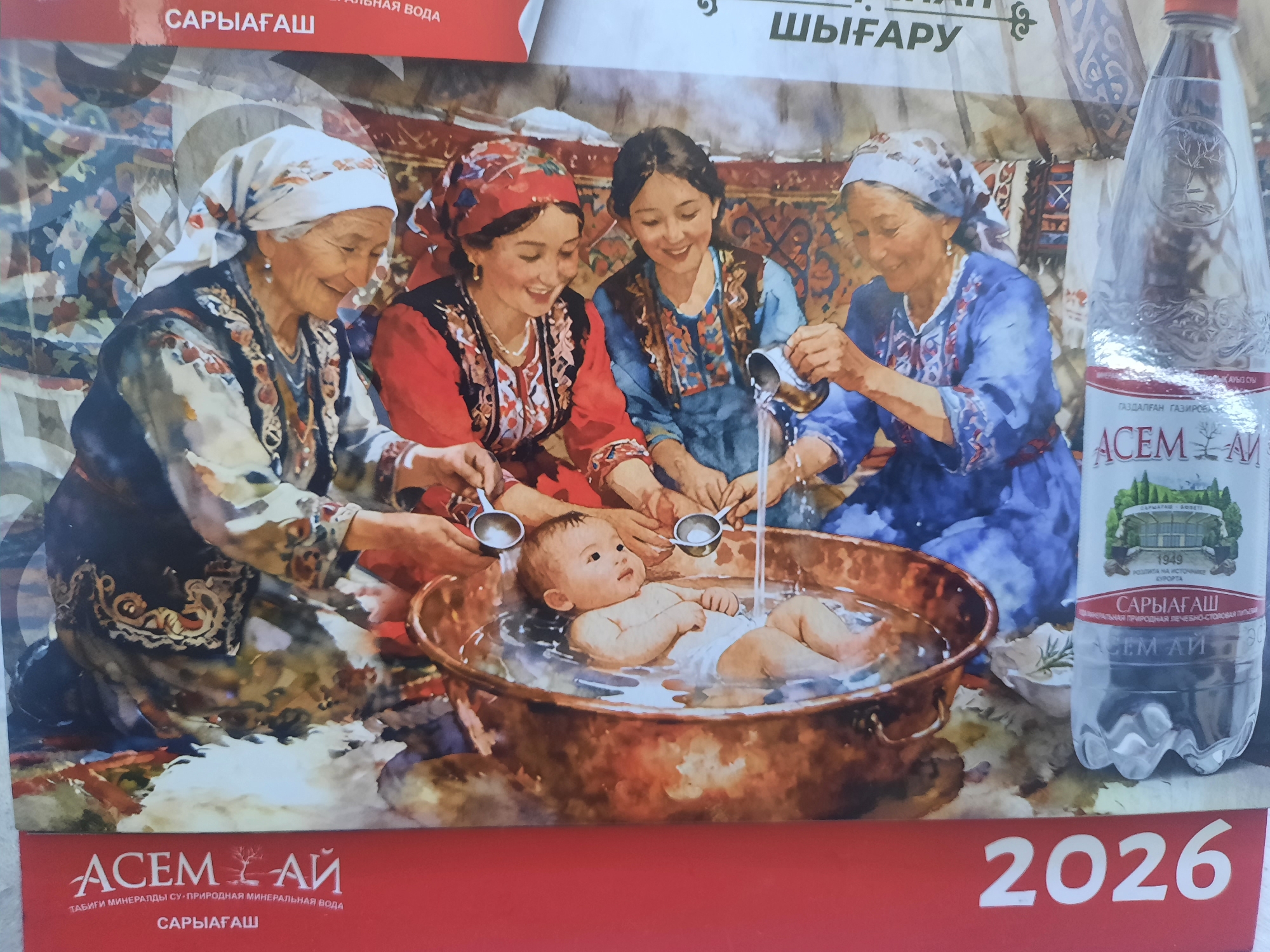 The image is a close-up, horizontally oriented photograph of a painting depicting a traditional scene. Four women, appearing to be of Central Asian descent, are gathered around a copper basin where a baby is being bathed. The women are dressed in traditional clothing with headscarves. Two older women on the left and right are wearing ornate, patterned garments and headscarves. Two younger women in the center are also wearing colorful attire and headscarves. One woman on the far right is pouring water into the basin from a small metal pitcher, while another woman to her left holds a similar pitcher. The baby is lying in the basin, which contains water and is surrounded by splashes. The background shows what appears to be a yurt interior with patterned textiles.
To the right of the scene, a bottle of "ACEM AI" mineral water is prominently featured. The label on the bottle displays text in Cyrillic and a crest with the year 1949. Below the painting, a red banner contains the "ACEM AI" logo and text that reads "ТАБИҒИ МИНЕРАЛДЫ СУ-ПРИРОДНАЯ МИНЕРАЛЬНАЯ ВОДА" and "САРЫАГАШ". The year "2026" is displayed in large white numbers on the right side of the banner. The location in the city of Zhanaozen, Kazakhstan, is not directly indicated within the image itself, but the branding and text on the bottle and banner suggest a connection to that region or a product originating from Kazakhstan.