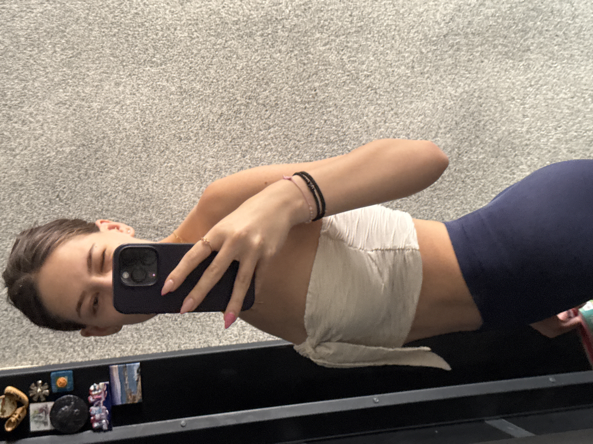The image is a selfie taken by a young woman in Sofia, Bulgaria. She is wearing a white, tie-front bandeau top and navy blue leggings. The woman is holding a dark-colored smartphone in her right hand to take the picture. She is winking and has a ring on her left ring finger, and two bracelets on her left wrist. Her nails are painted bright pink.
The background is a textured gray wall, possibly carpet or a similar fabric. Below the woman, there is a black surface with several small objects, resembling decorative pins or magnets, arranged on it. These objects include a gold-colored item shaped like a flower, a small blue square with an orange circle, a black circular object, a colorful miniature figurine of a bear-like creature, and a small rectangular picture of a landscape.
The lighting suggests it could be daytime indoors, with no indication of specific weather conditions. The overall mood seems casual and playful due to the woman's pose and facial expression.
