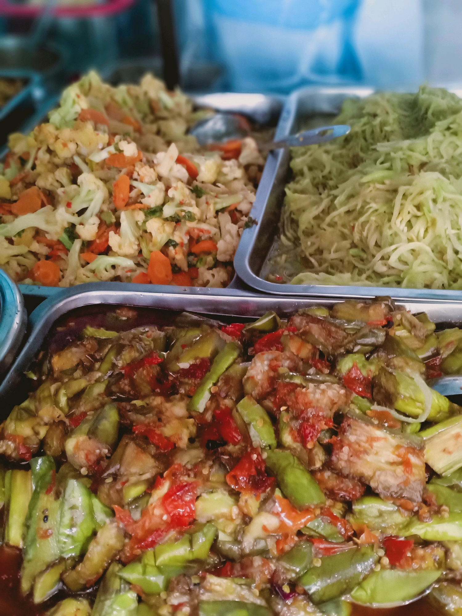 In the city of Sumberrejo, Indonesia, a close-up, overhead shot captures three silver metal trays filled with various dishes, likely from a food stall. The foreground is dominated by a tray of stir-fried eggplant with chilies, its pieces cut into irregular lengths and coated in a rich, reddish-brown sauce. Interspersed are visible slivers of red chili and some darker, possibly fried, eggplant pieces. Behind this, a tray to the left contains a medley of cooked vegetables, primarily cauliflower florets and sliced carrots, with hints of green vegetables and a reddish spice. The furthest tray on the right holds a mound of finely shredded green vegetables, possibly cabbage or papaya, appearing light and fresh. The overall impression is one of diverse, prepared food ready for serving.