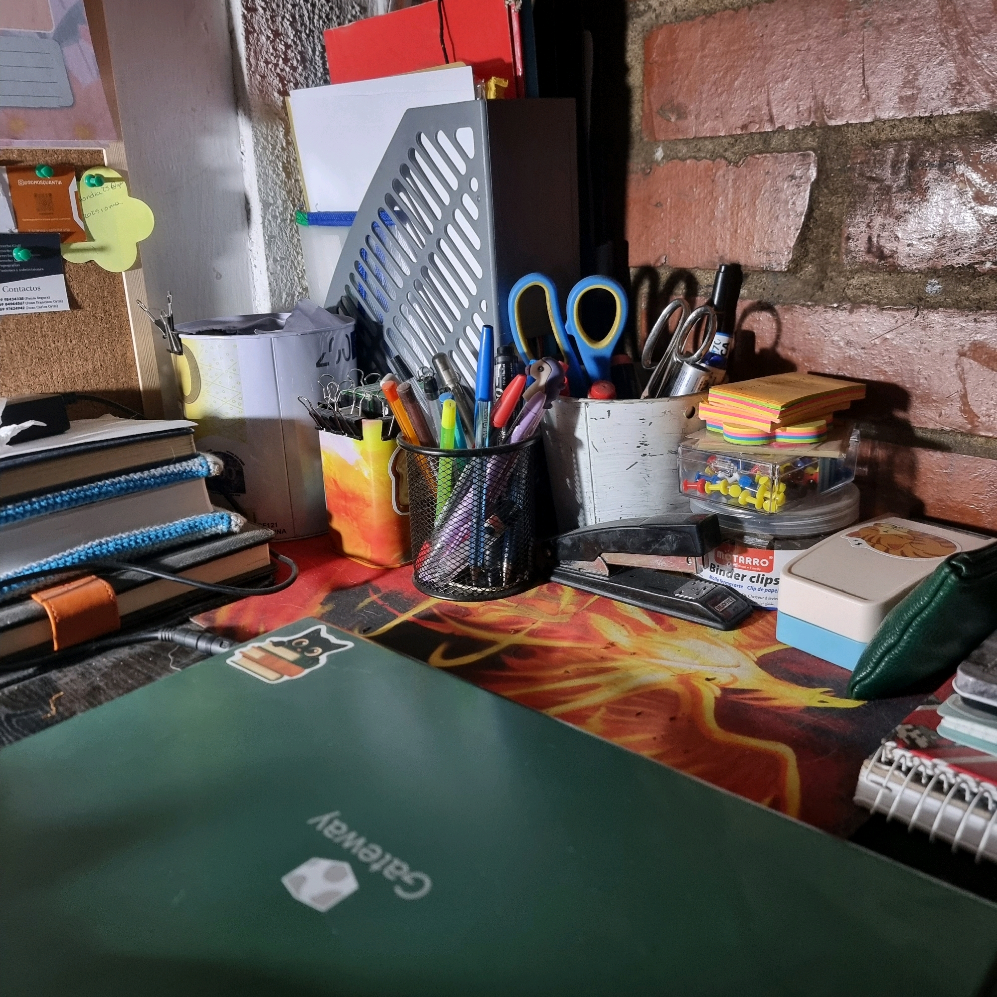 The image displays a desk cluttered with various office supplies and personal items. A green laptop is prominent in the foreground, featuring a sticker of a cat reading a book. To the right of the laptop, a stapler sits on a desk mat with a fiery graphic. Several containers hold pens, pencils, and scissors. Stacks of notebooks and small colorful sticky notes are also visible. A corkboard with notes and thumbtacks is positioned on the left side of the image. The background reveals a brick wall and part of a white shelf unit. The scene appears to be an indoor workspace, possibly a home office or study area. The specific location in Rengo, Chile, is not determinable from the visual information provided in the image.