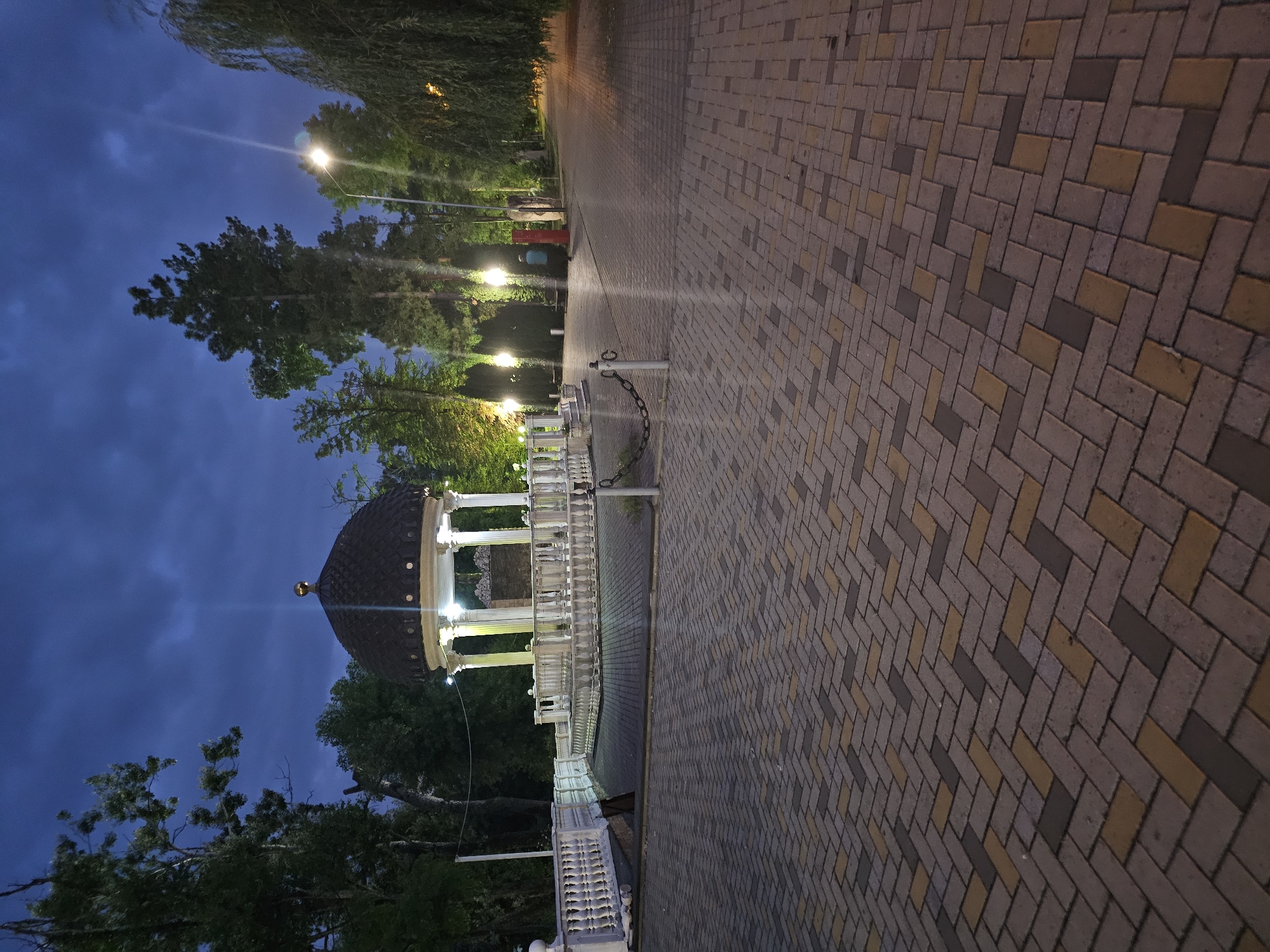 An image taken at dusk in Cherkessk, Russia, depicts a park with a paved walkway and a gazebo. The sky is a deep blue with some clouds. The gazebo, a white structure with a dark, domed roof, is illuminated from within, casting a warm glow. It is surrounded by a white, ornate balustrade. The walkway in the foreground is paved with interlocking rectangular bricks in shades of gray, yellow, and brown, laid in a herringbone pattern. Trees line the walkway, some with lush green foliage, while others appear more sparse. Several lampposts with bright lights are visible along the path, illuminating the area and creating streaks of light in the image. A chain link fence with a white post is present near the gazebo. In the distance, a lone figure in a blue shirt and dark pants walks away from the camera down the path. The overall scene suggests a quiet evening in a public park.