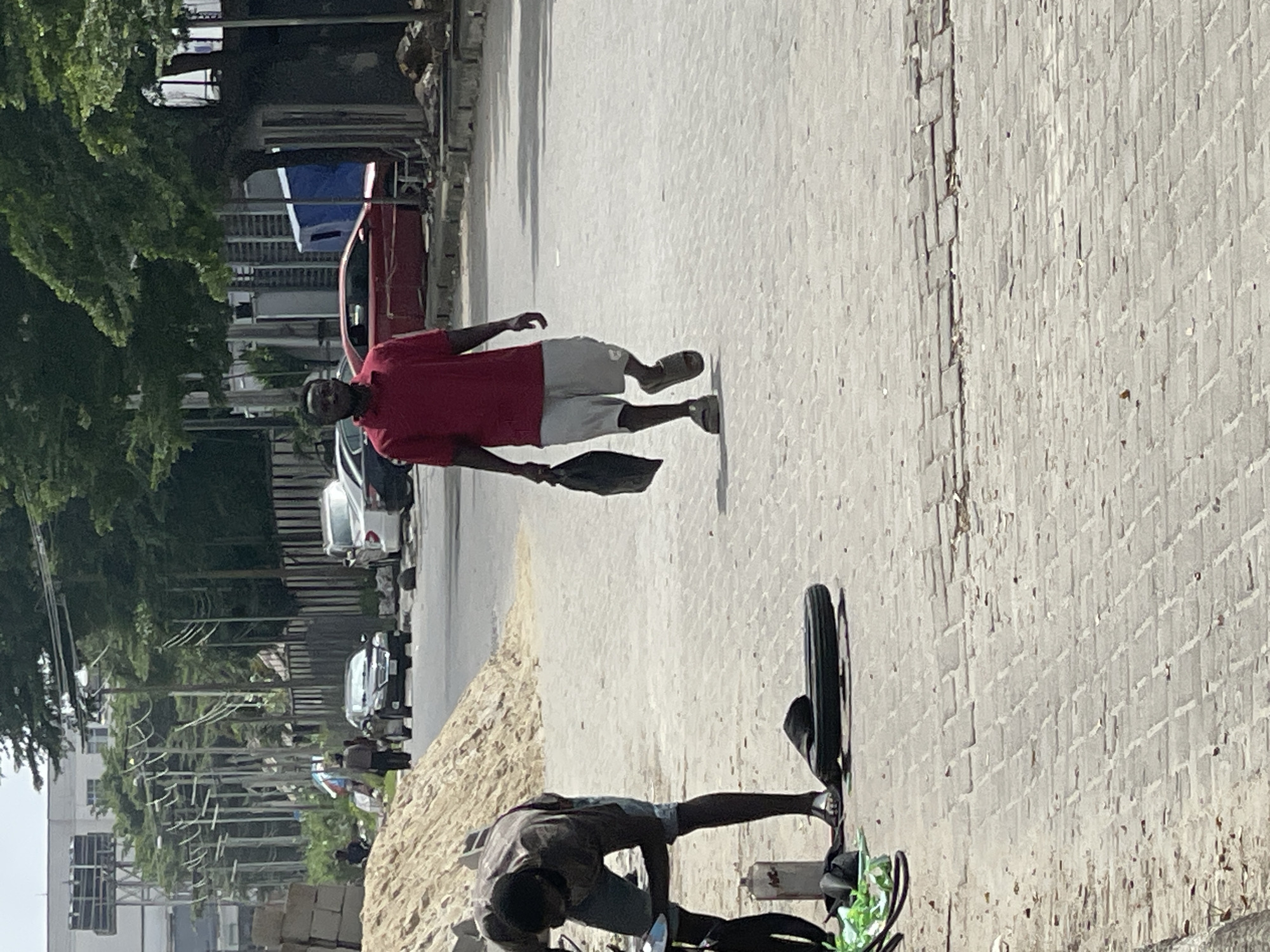 In Igboefon, Nigeria, on a sun-drenched street paved with interlocking bricks, a man in a red polo shirt and grey shorts walks with a black plastic bag in his right hand. He is wearing brown sandals. To his left, another man, wearing a brown t-shirt and denim shorts, is bent over, working on what appears to be a tire and a bottle of liquid on the ground. Green plastic items are also visible near him. In the background, a large pile of sand sits beside the road, with construction materials and several parked cars, including a white SUV and a red sedan, on the street. Trees line the background, and utility poles with numerous wires are visible.