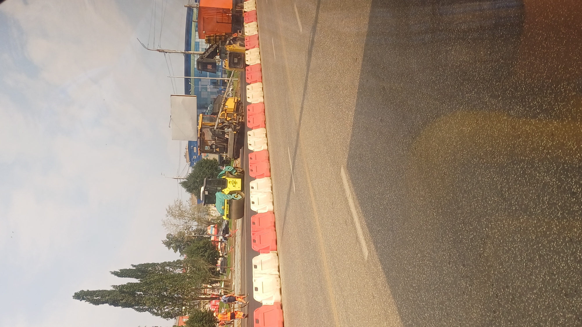 The image depicts a street scene in Izhevsk, Russia, during daylight. The foreground shows a paved road with white and red plastic barriers delineating a construction area. In the background, several pieces of construction equipment are visible, including a road roller, an asphalt paver, and a truck with a crane. Workers in high-visibility vests are present, engaged in road work. The surrounding environment includes trees, buildings, and a cloudy sky. The overall impression is one of active road construction and maintenance.
