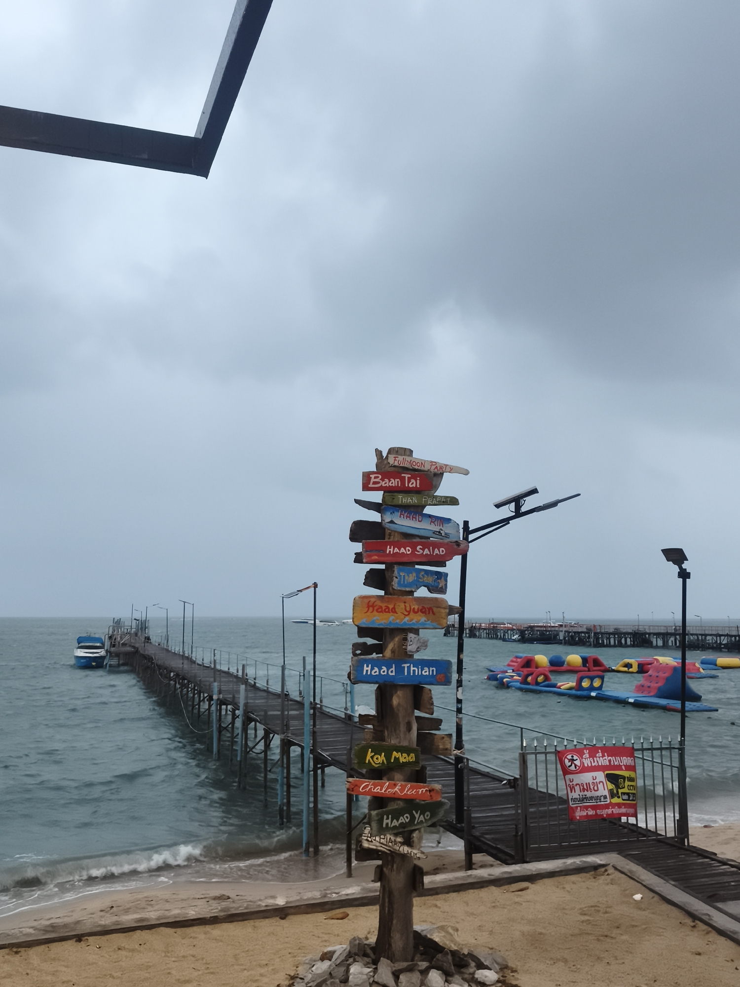 This is a medium-shot, eye-level, outdoor, daylight photograph taken on the island of Ko Samui, Thailand. The scene depicts a weathered wooden pier extending into a choppy, gray sea under an overcast sky. On the beach in the foreground, a signpost with multiple directional signs, pointing to various locations including "FullMoon Party", "Baan Tai", "Haad Rin", "Haad Salad", "Haad Yuan", "Haad Thian", "Koh Maa", "Chaloklum", and "Haad Yao", stands prominently. The signs are colorful but faded, indicating age and exposure to the elements. To the left of the pier, a small blue and white boat is moored. In the mid-ground, a floating water park with inflatable slides and obstacles is visible. A metal gate with a red sign in Thai, warning about unauthorized entry and mentioning CCTV, is situated on the right side of the image, near the entrance to the pier. The beach itself is sandy with scattered rocks and debris. The overall mood of the image is somber, likely due to the cloudy weather. No people are visible in the image.