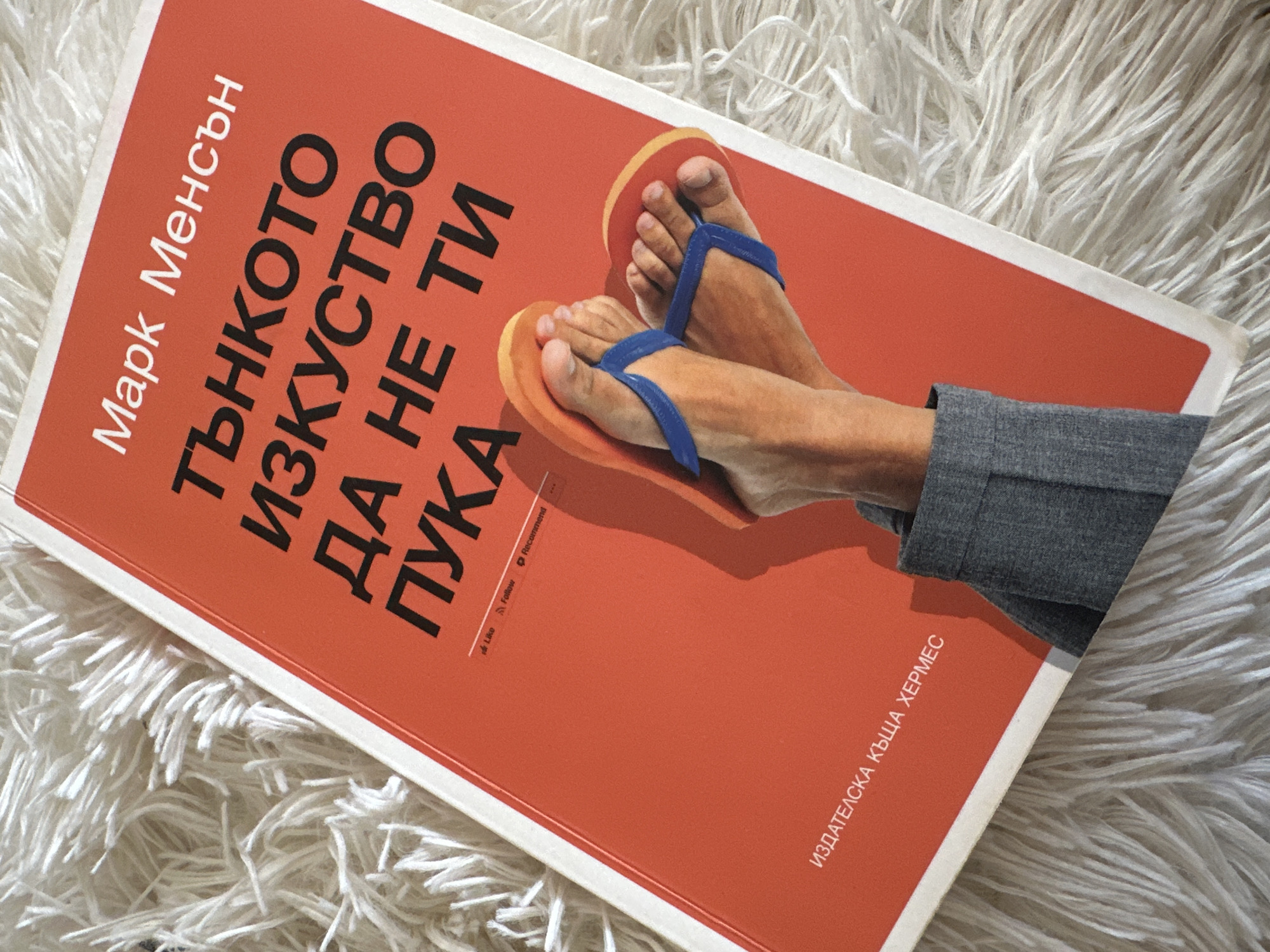 A rectangular book with a vibrant orange cover lies diagonally on a white, shaggy, fuzzy surface, resembling a rug or blanket. The book's cover features an illustration of a pair of human feet, clad in orange and yellow flip-flops with blue straps. The feet appear to be extending from a grey-sleeved garment, suggesting a relaxed posture.
The text on the book cover is in Bulgarian. The author's name, "Марк Менсън" (Mark Manson), is printed vertically in white on the left side. The title, "Тънкото изкуство да не ти пука" (The Subtle Art of Not Giving a F*ck), is prominently displayed in black, large font. Towards the bottom right, also printed vertically, is the publisher's name: "ИЗДАТЕЛСКА КЪЩА ХЕРМЕС" (HERMES PUBLISHING HOUSE). Small, almost unreadable icons and text, possibly related to social media ("Like", "Follow", "Recommend"), are visible near the spine.
The scene depicts the book in a casual indoor setting, emphasizing its theme of nonchalance and practicality, aligning with the implied content of the book. The image is set in Sofia, Bulgaria.