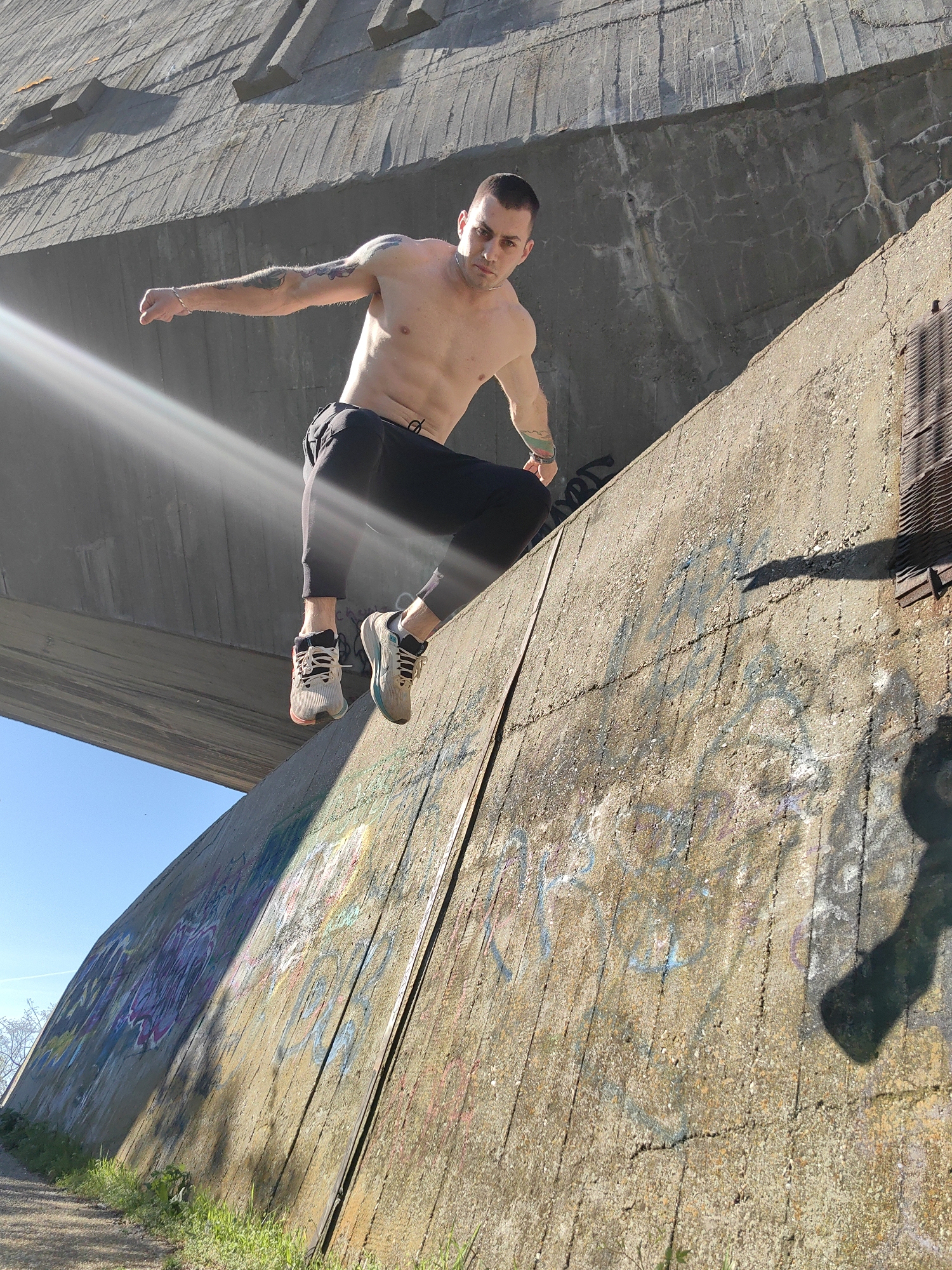 A dynamic, wide-angle photograph captures a muscular man performing an acrobatic maneuver on a large concrete wall under a clear blue sky in Varna, Bulgaria. The shirtless man has short, dark hair, prominent arm tattoos, and a silver bracelet on his right wrist. He wears dark athletic pants and two-toned sneakers, one with pink/orange accents and the other with blue/teal. He is caught in a moment of athletic exertion, with his body angled, one foot firmly planted on the steep concrete wall and the other slightly lifted, as if pushing off or maintaining balance. His arms are outstretched, and his left hand is clenched into a fist. His expression is focused and intense, looking directly towards the camera.
The scene is dominated by massive concrete structures, suggesting an urban or industrial setting. A large, smooth concrete bridge or underpass spans diagonally across the upper part of the frame, casting a shadow below. The concrete wall the man is on is textured, with vertical grooves and signs of wear, and is extensively covered in colorful, abstract graffiti. At the base of the wall, green grass and sparse vegetation are visible along a dirt path. Strong sunlight from the left creates a bright lens flare and casts distinct shadows across the scene, indicating midday or early afternoon. A metal grate or vent is visible on the upper right section of the wall. The image conveys a sense of energy and skill, capturing a fleeting moment of parkour or free-running against a rugged, sunlit backdrop.