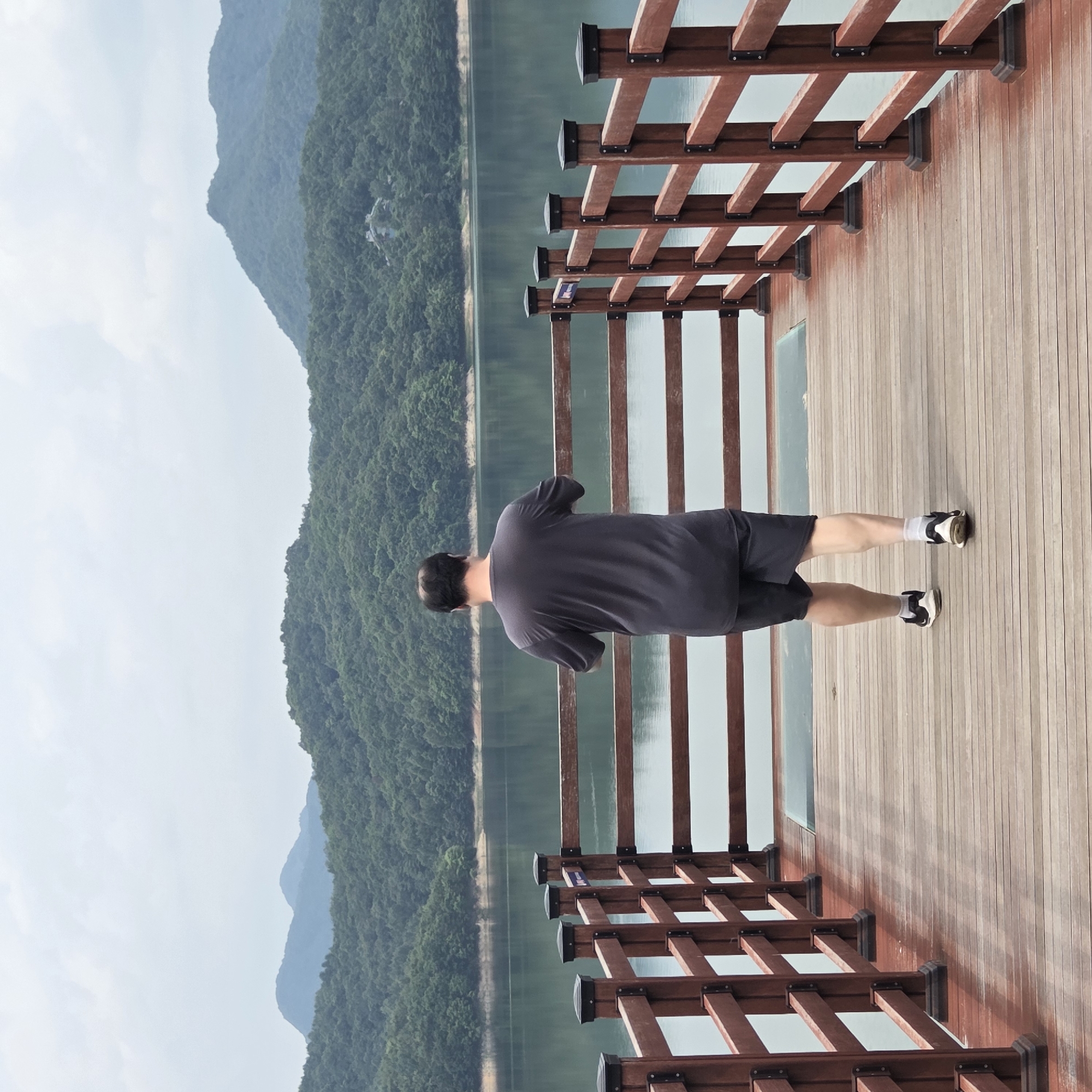 A man with short, dark hair, seen from behind, stands on a wooden boardwalk. He wears a dark grey t-shirt and dark shorts. His legs are bare below the shorts, and he is wearing white socks and black athletic shoes. The boardwalk has a railing made of dark red wooden posts and horizontal planks, with black decorative caps on the posts. The boardwalk extends forward and to the right of the frame. In the background, a large body of water with a greenish hue is visible, reflecting the overcast sky. Beyond the water, a densely forested hillside rises, topped by hazy, blue-green mountains under a cloudy sky. A small, light-colored building with a distinctive roofline is partially visible nestled on the hillside. The scene appears to be an outdoor recreational area, possibly a park or a scenic viewpoint, located in Gapcheon-myeon, South Korea.