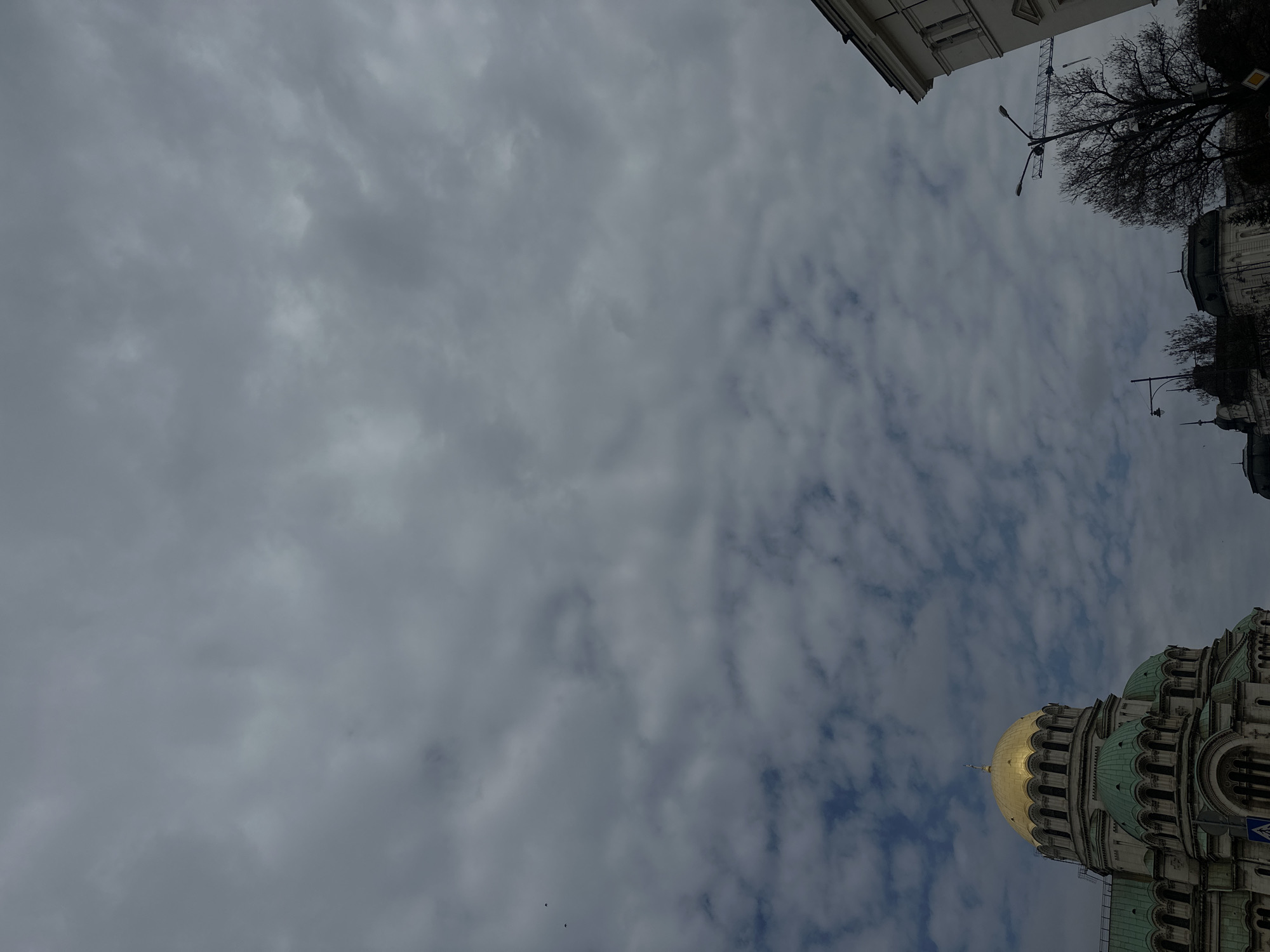 The image shows a view of the Alexander Nevsky Cathedral in Sofia, Bulgaria, from a low angle, looking upwards. The sky is mostly overcast with patches of blue. The cathedral's golden dome is visible. Parts of the cathedral's architecture, including green-colored details and windows, are also seen. Adjacent buildings and trees are partially visible. A street lamp stands next to a nearby building.