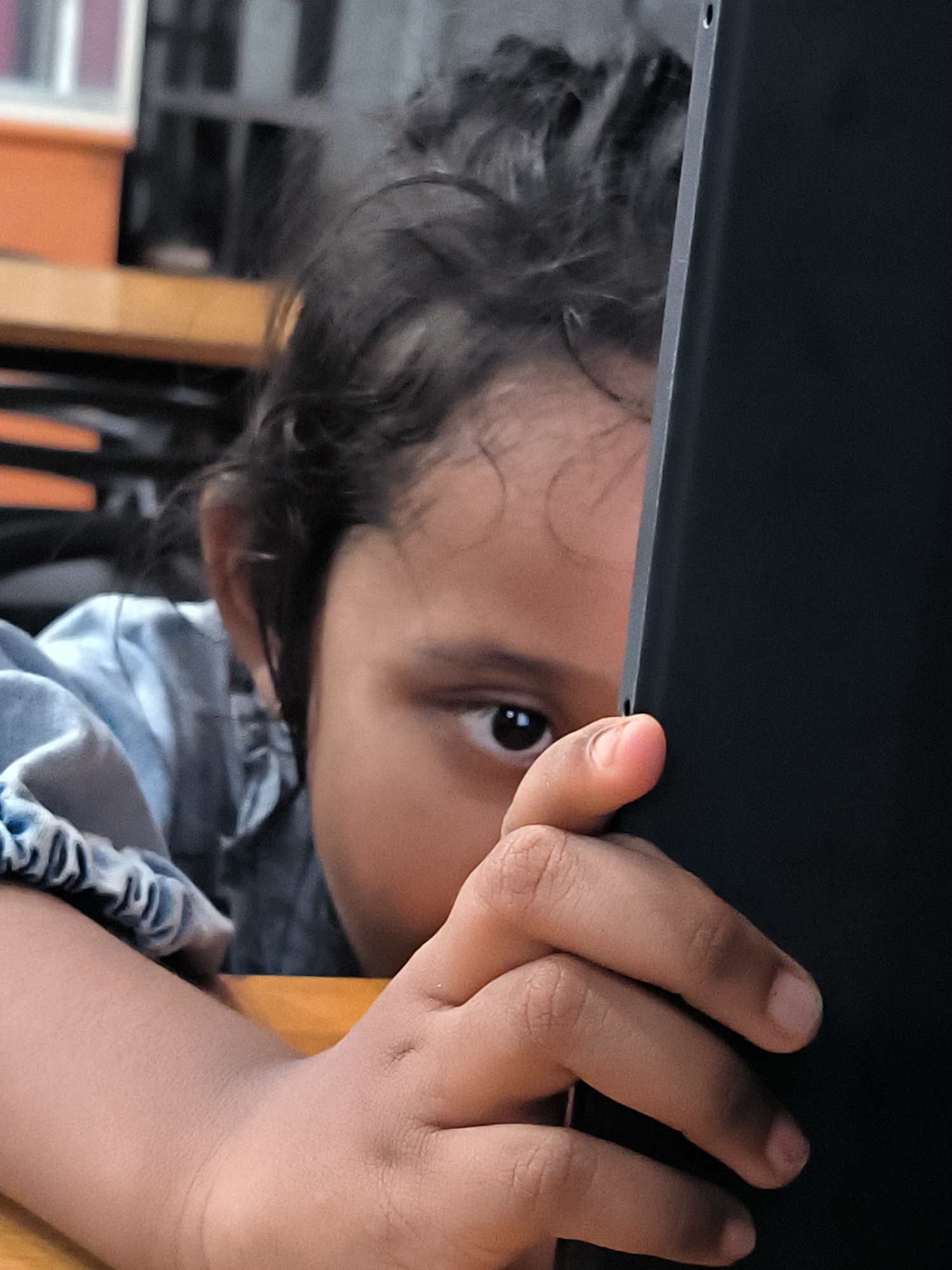 A young child is partially obscured by a dark rectangular object, possibly a tablet, held in their hand. The child's left eye is visible, gazing forward. The child has dark, curly hair. They are wearing a light blue shirt. The child's left arm rests on a wooden surface. The background is blurry, but hints at an interior setting with orange and black elements, suggesting furniture or decor. The image shows an intimate moment, perhaps the child is absorbed in the screen. The specific location within Lhokseumawe, Indonesia, is not discernible from the image.