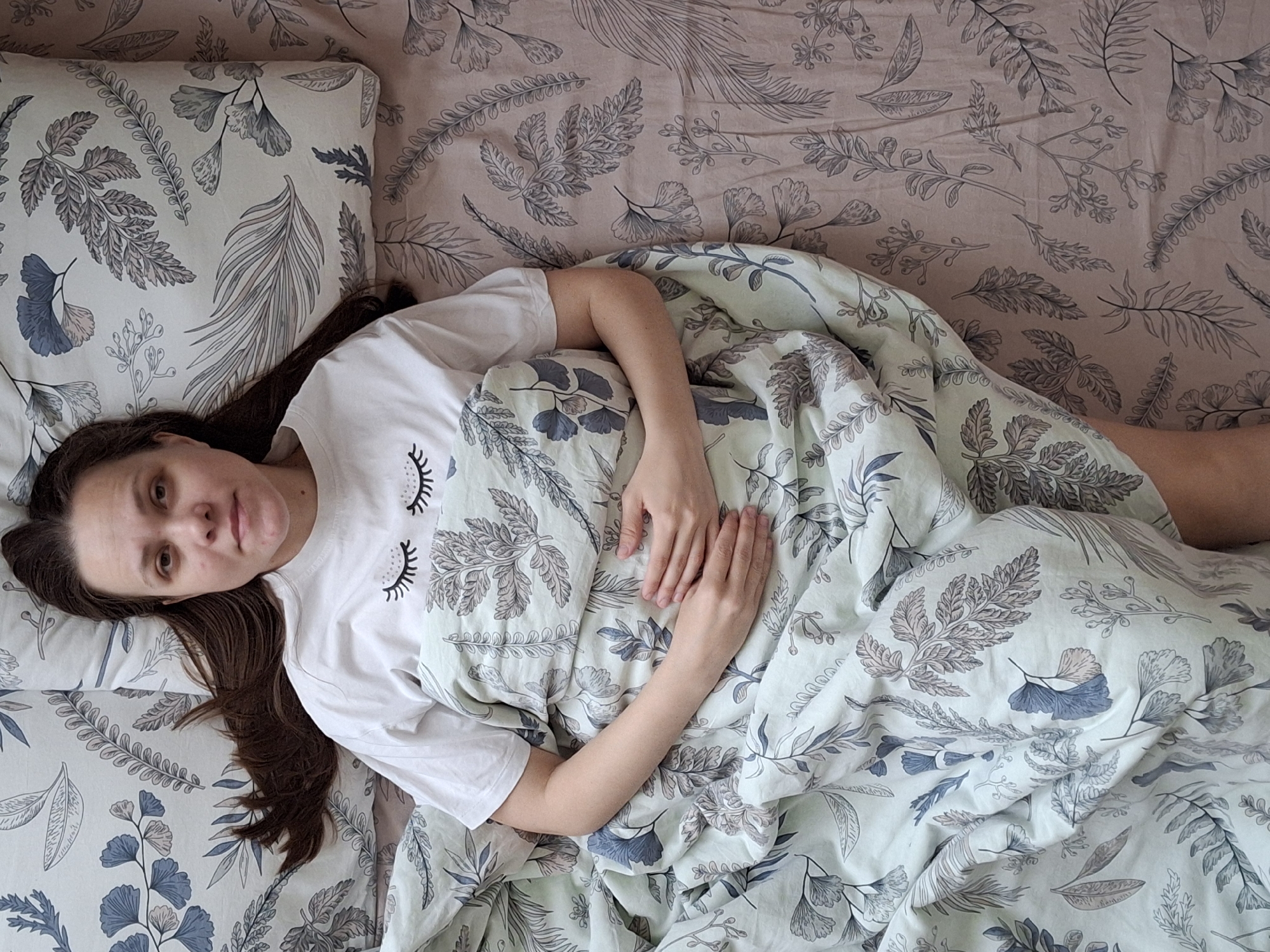 A woman is lying in bed, partially covered by a floral-patterned duvet. She wears a white T-shirt with an image of eyelashes. The bed has a floral-patterned pillow and bedding. The location is Chaikovsky, Russia.