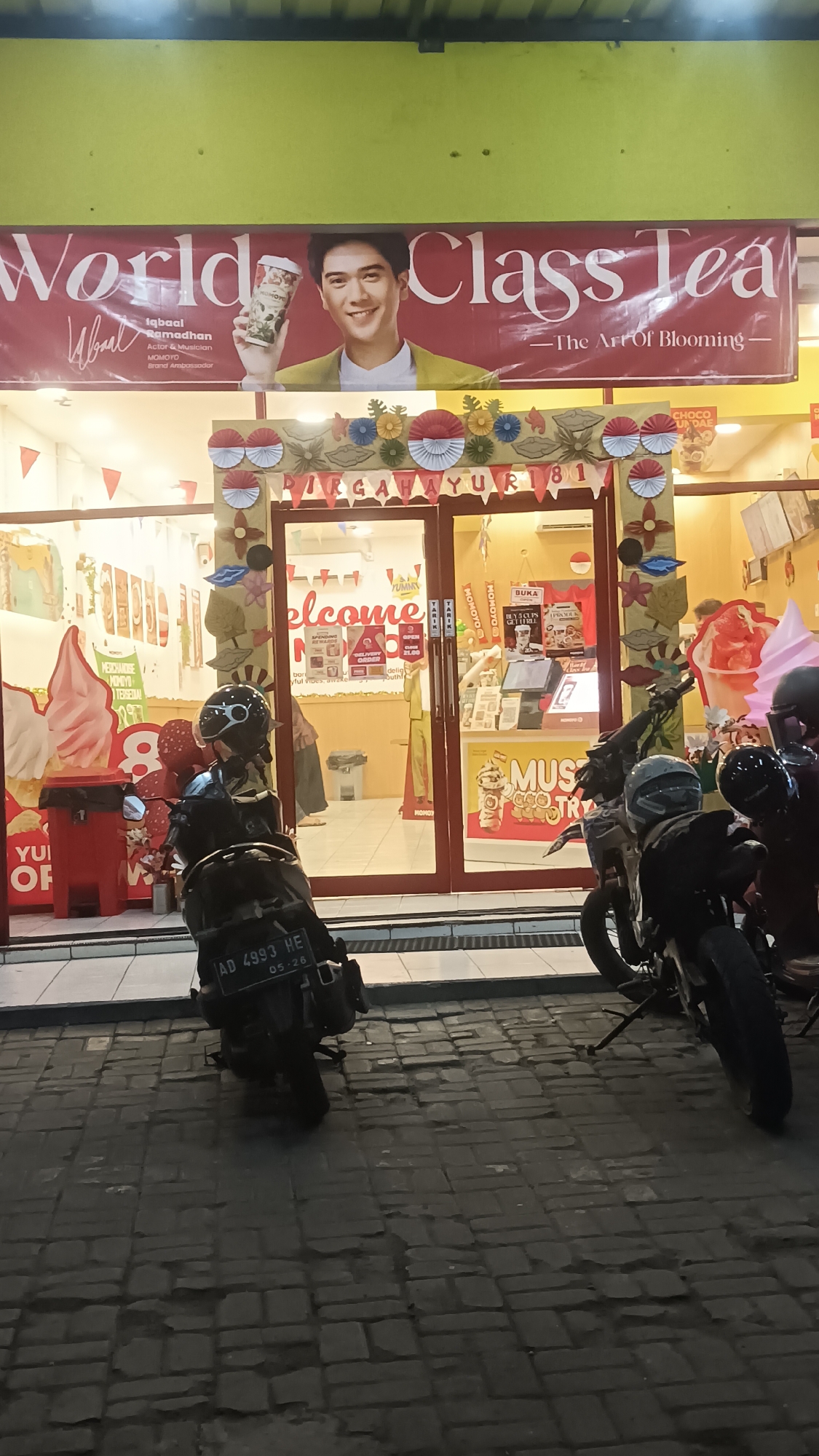 This is a medium shot, taken at night, of the exterior of a Momoyo bubble tea shop, likely located in the city of Sragen, Indonesia. The shop's facade is brightly lit, with a large red banner across the top featuring white text that reads "World Class Tea". Below the banner, a younger East Asian male, identified as Iqbal Ramadhan, actor and musician, and MOMOYO brand ambassador, is pictured holding a cup. Decorative banners featuring paper fans in red and white, a motif commonly associated with Indonesian independence day, are hung above the shop entrance. The shop's entrance is a double glass door, with "welcome" in large red letters visible inside. Displays for bubble tea and ice cream are visible through the glass. In front of the shop, several motorcycles are parked on a brick-paved sidewalk. One motorcycle in the foreground has its license plate clearly visible: AD 4993 HE, with the expiration date 05-26. The overall impression is a commercial establishment at night, with elements of local culture and national pride evident in the decorations.