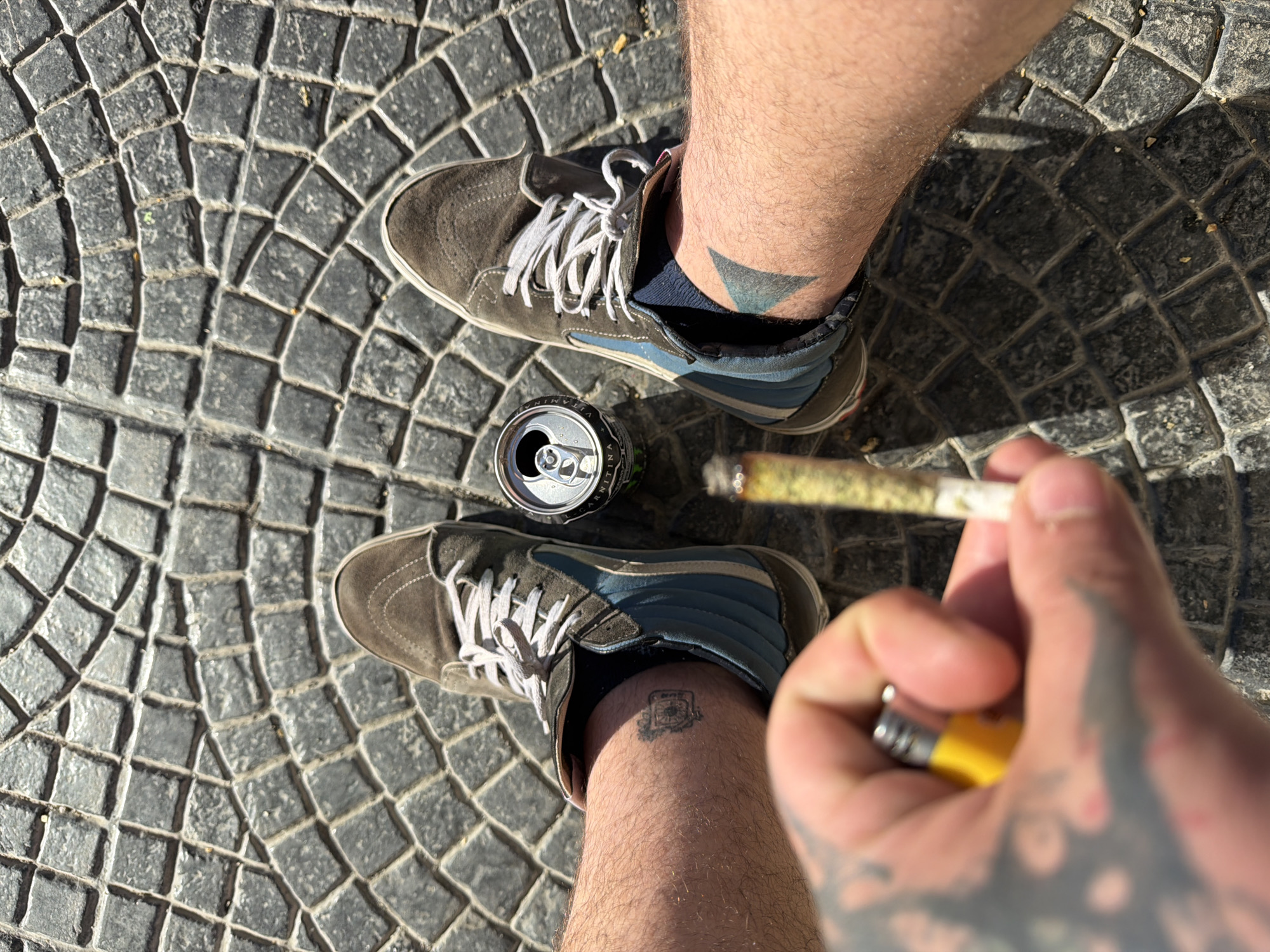 The photograph is taken from an overhead perspective, capturing a scene occurring outdoors on a paved surface. The paving consists of small, dark gray cobblestones arranged in a circular pattern. A pair of legs and feet, wearing tan Vans-style sneakers with blue accents and white laces, is positioned in the center of the frame. The right leg features a tattoo on the inner ankle, while the left leg has a triangle-shaped tattoo. In the space between the two legs, an open aluminum can is visible.
The right hand is holding a lit cigarette and a yellow lighter. Tattoos cover the hand and part of the forearm.
No information is available from the provided image confirming that the image was taken in Ushuaia, Argentina.