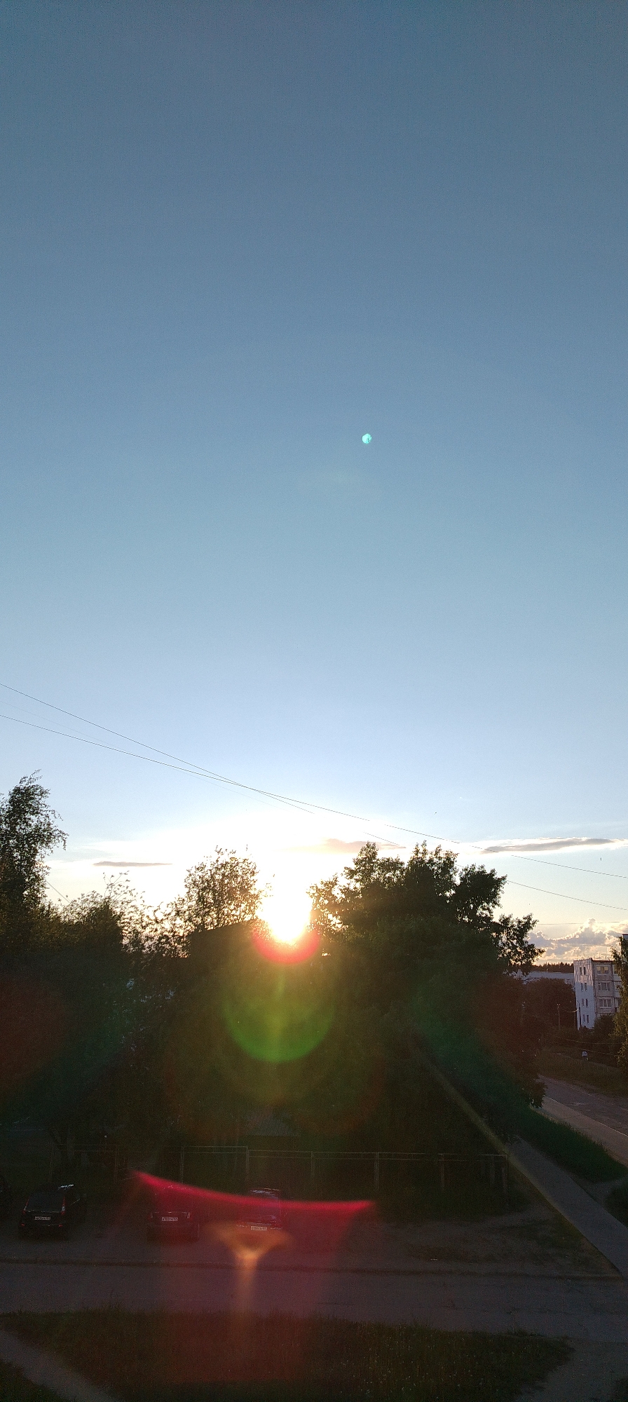 The image captures an outdoor scene at what appears to be sunset, looking out from an elevated perspective over a residential or suburban area. The sun is low on the horizon, partially obscured by a dense line of trees, and directly facing the camera, creating significant lens flare with prominent red and green circular halos and streaking across the lower half of the image.
The upper portion of the frame is dominated by a clear, light blue sky, which transitions to a brighter, almost white hue near the horizon where the sun is setting. A distinct, bright, circular, slightly greenish-blue artifact, likely a lens reflection, is visible in the upper center of the sky. A few thin, wispy clouds are present along the horizon, primarily to the right of the sun. Several dark, thin power lines stretch across the middle of the sky above the trees.
The middle ground is filled with lush, dark green foliage from numerous trees, silhouetted against the bright sun. Beyond the trees on the right, a multi-story white building with visible windows can be seen in the distance, suggesting a populated area.
In the foreground, a paved surface, resembling a street or parking lot, is visible. Two dark cars are parked on this surface on the left side of the frame. One car has a visible Russian license plate, "A 907 EH 60", and another has "A 823 TM 60", indicating the Pskov Oblast region, which is a notable detail considering the provided location context of Chaikovsky, Russia. A low fence or barrier runs along the edge of the paved area, separating it from the wooded area. The overall mood is serene, capturing the golden hour of the day.