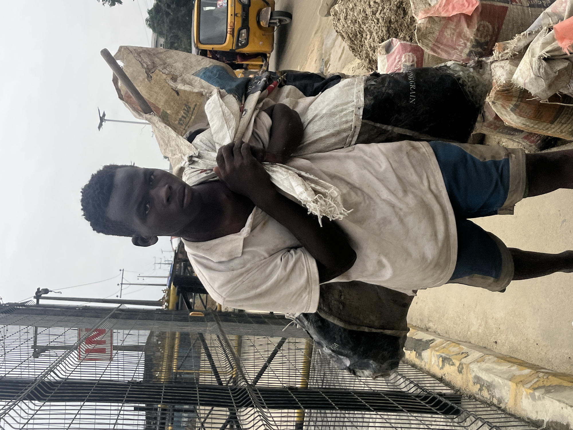 A young dark-skinned man stands in the city of Ado, Nigeria, carrying a large sack of materials over his shoulder. He wears a light-colored polo shirt and blue shorts. The sack is made of rough material and appears to be supported by a wooden pole. Behind him, a yellow three-wheeled vehicle, commonly known as a Keke Napep, is visible, along with piles of sacks filled with what looks like sand or cement. A metal fence runs along the left side of the frame, with a red and white sign attached to it. The ground is paved, and the sky is overcast. The overall scene suggests a work or transportation context in a bustling urban environment.