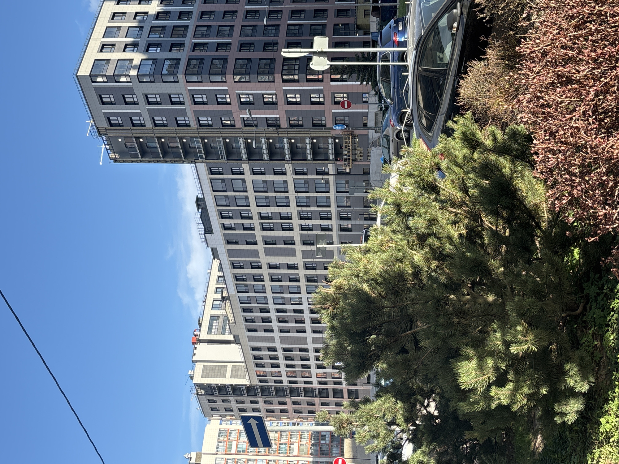 This media file captures a vibrant urban street scene in Saint Petersburg, Russia, under a clear, bright blue sky. The image is composed of modern multi-story buildings, parked cars, and lush vegetation, suggesting a sunny daytime setting.
In the foreground and midground on the right, a large, dark green coniferous tree, likely a pine, dominates the view, its branches partially obscuring other elements. Adjacent to it, a bush with dense reddish-brown foliage adds a contrasting texture. Behind this greenery, several cars are parked alongside what appears to be a street or parking area. A blue sedan and a dark-colored vehicle are clearly visible.
The background is dominated by contemporary high-rise buildings. The most prominent structure features a sleek, modern design with a facade patterned in shades of grey, dark grey, and light beige panels, punctuated by numerous large windows. A distinctive architectural detail includes a multi-story cantilevered section that extends outwards from the main building. Further in the background, another building with a different aesthetic, featuring more traditional orange or reddish-brown elements, can be seen.
Traffic signs are visible, including a blue rectangular sign with a white upward-pointing arrow, typically indicating "straight ahead" or "one way," and a red circular sign with a white horizontal bar, signifying "No Entry" or "Do Not Enter." A street light pole is also present. The clear blue sky with a few faint clouds suggests fair weather. A black power line or cable runs diagonally across the upper left portion of the frame. No people or specific activities are depicted; the scene is a static snapshot of an urban environment.