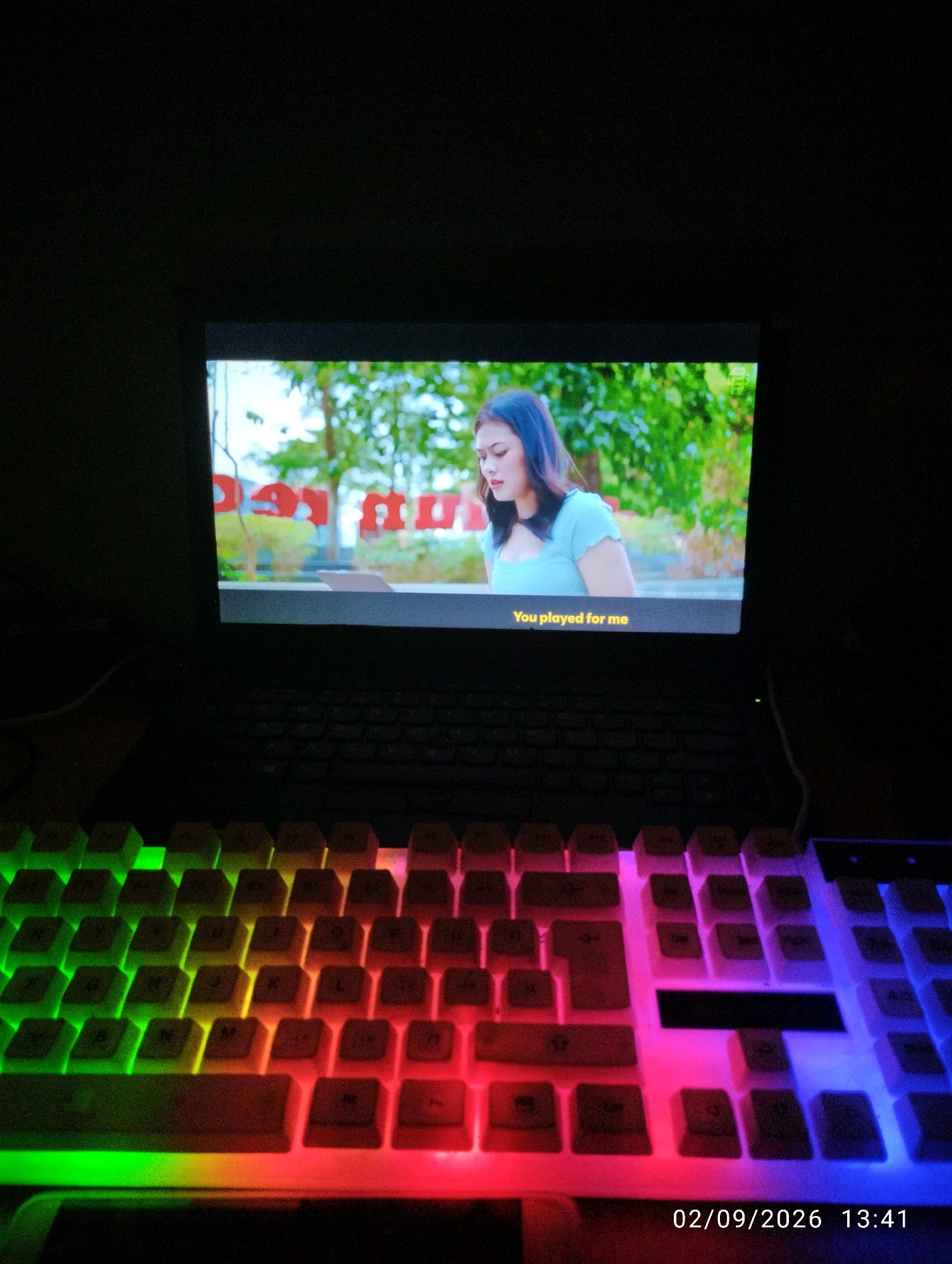 In a dimly lit room, a laptop displays a scene with a woman and text overlay reading "You played for me." In the foreground, a keyboard with customizable RGB lighting illuminates the scene with a gradient of green, yellow, red, pink, and blue hues. The overall setting appears to be a personal space, likely a home office or entertainment area, located within the city of Purworejo, Indonesia. The presence of a laptop and keyboard suggests activities related to computing or gaming. The timestamp "02/09/2026 13:41" indicates the time and date the image was captured.