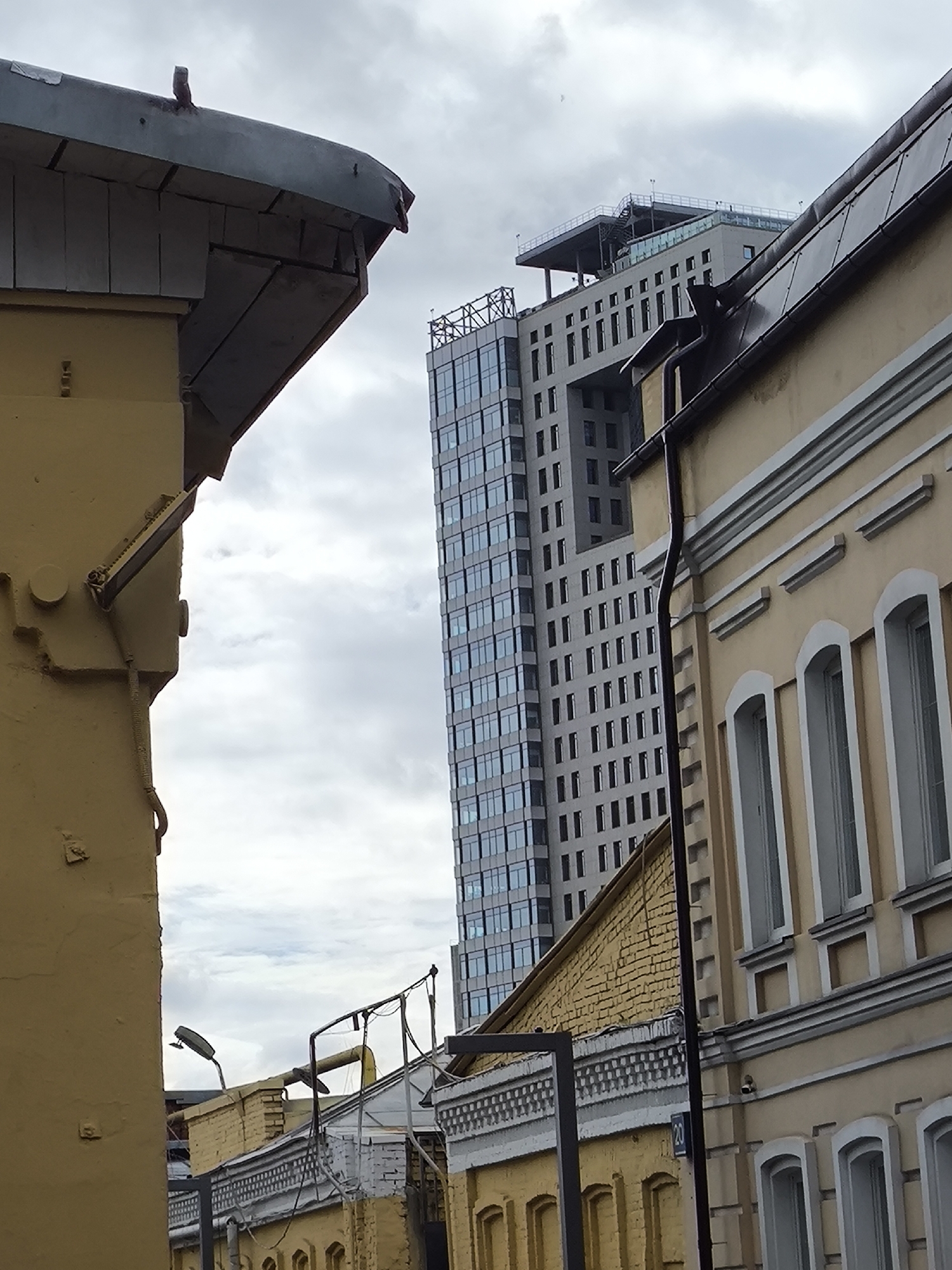 The image shows a street scene in Moscow, Russia. An old, yellow-painted building is in the foreground, partially obstructing the view of a modern, tall skyscraper with a flat rooftop. A smaller, yellow-brick building with arched windows is visible between the older building and the skyscraper, with streetlights and wires above it. The sky is overcast with gray clouds.