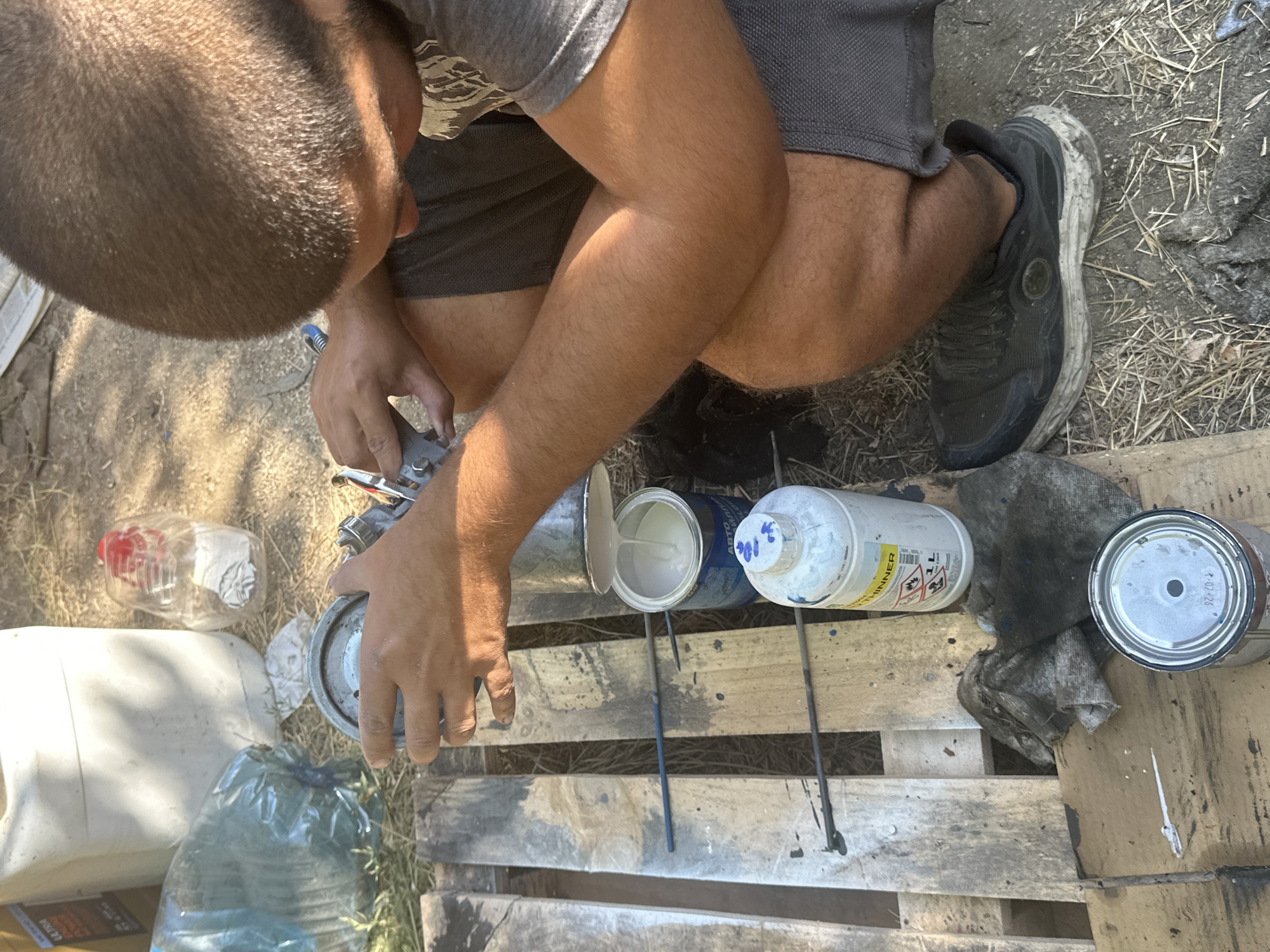 In Galiche, Bulgaria, a man is crouched on a wooden pallet, engaged in what appears to be a painting or cleaning activity. He is holding a paint can upside down, pouring white liquid into another open paint can below. A spray gun is also visible in his hands, suggesting he might be using it in conjunction with the paint. Surrounding the pallet are various items including a large white plastic container, a clear plastic bottle with a red cap, a green plastic bag, a white bottle labeled "THINNER," a dirty rag, and another smaller paint can on a piece of cardboard. The ground is dirt and dry grass, indicating an outdoor, possibly rural setting. The man is wearing a grey t-shirt and dark shorts, and a dark athletic shoe. The lighting suggests it is daytime, with shadows cast by the objects and the man.