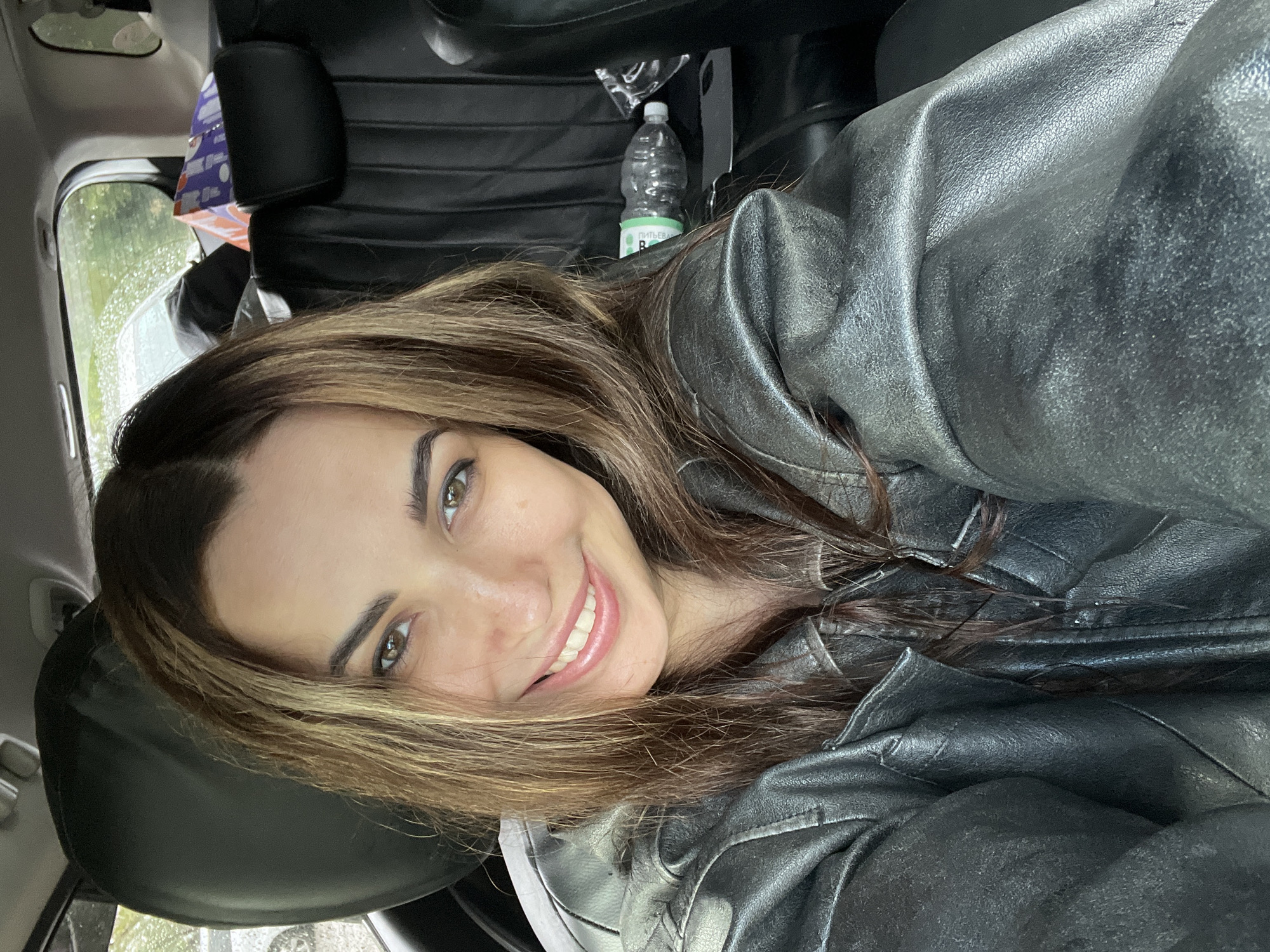 The image, oriented with the woman's head to the left, captures a selfie of a young woman smiling inside a car. She has long, dark brown hair with prominent lighter, possibly caramel, highlights that frame her face and cascade over her shoulders. Her eyes are brown or hazel, and she displays a wide, cheerful smile showing white teeth. She is wearing a textured, dark, possibly metallic-grey or black leather/faux-leather jacket.
The setting appears to be the interior of a car. Black upholstered car seats and headrests are visible in the background. To the right of her head, a plastic water bottle with a green and white label is visible, on which Russian text "ПИТЬЕВАЯ ВОДА" (Drinking Water) and a partial letter "B" or number "8" can be discerned. Through the car window in the upper left, rain is visible on the glass, indicating wet weather outside. A patterned bag or package, with purple and white colors, is also partially visible behind a headrest. The lighting suggests it is daytime, and the woman's expression conveys happiness or contentment.