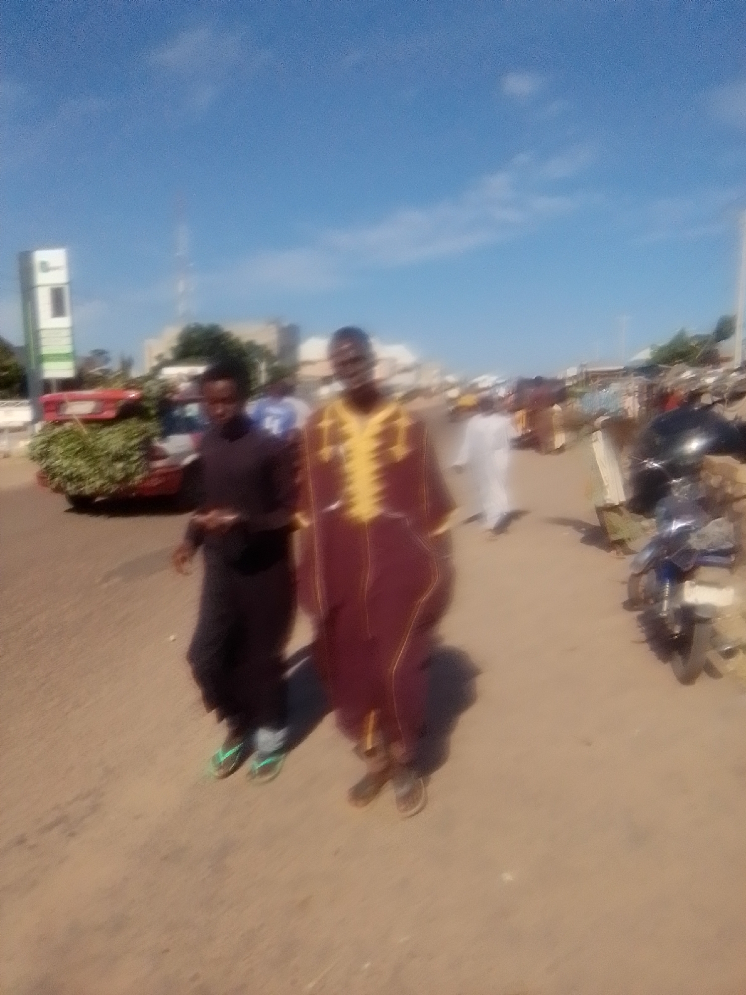The image depicts an outdoor scene, likely a street in Pantame, Nigeria, under a clear blue sky with some wispy clouds. In the foreground, two men are walking. The man on the left is wearing a dark, possibly black, outfit and light-colored sandals. The man on the right is wearing a traditional maroon garment with yellow intricate embroidery around the neck and chest, and he is also wearing sandals. To the left of the men, a red vehicle is partially visible, loaded with green foliage. Further in the background, there are structures that appear to be shops or buildings, and a tall white tower with green accents, possibly a communication tower or landmark. On the right side of the image, a black motorcycle is parked. Other individuals and activities are blurred in the background, suggesting a busy street or marketplace environment. The ground is a sandy, earthy color.
