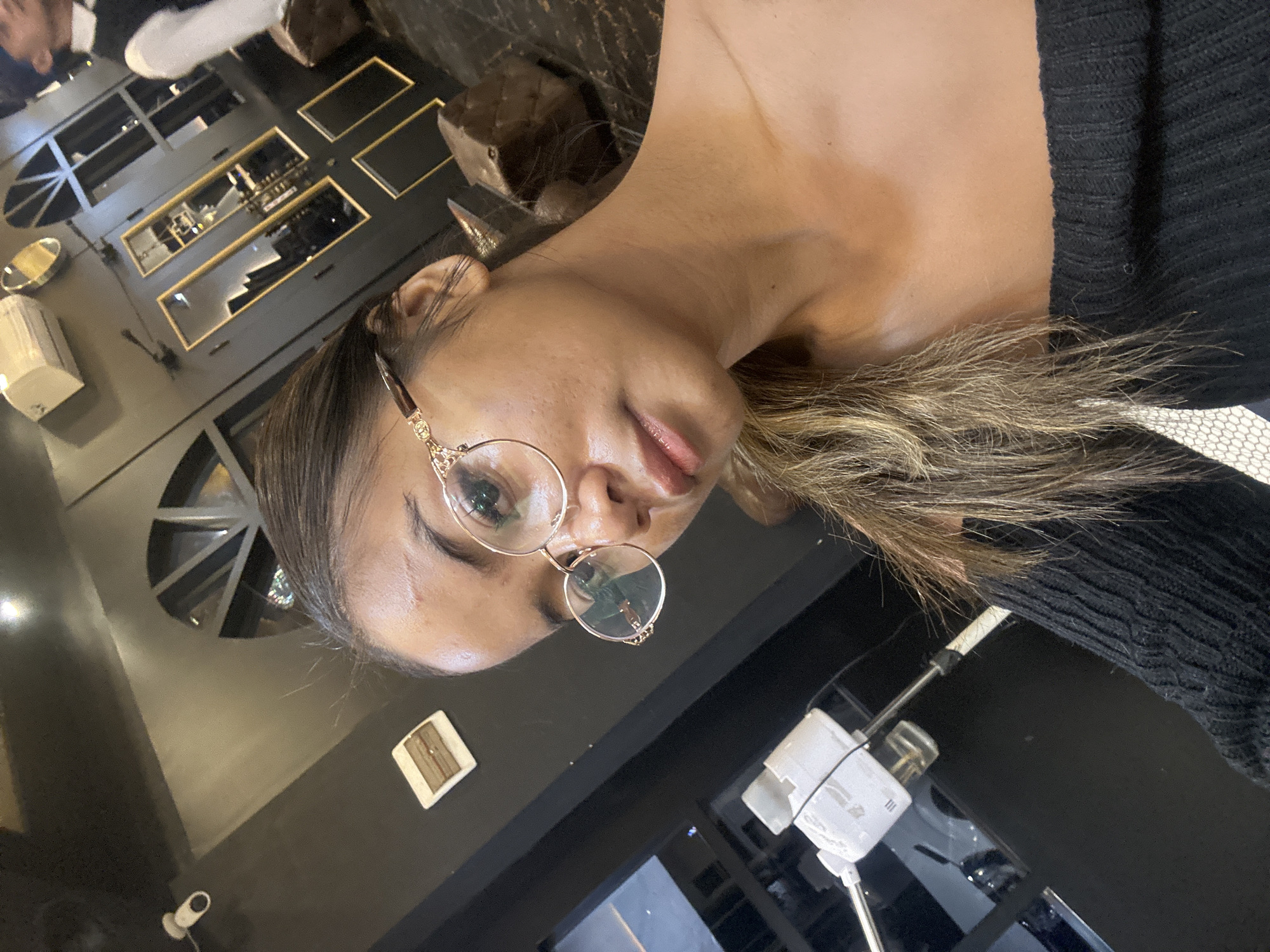 This image, taken in North Jakarta, Indonesia, presents a medium close-up of a woman looking directly at the camera. She has medium-brown hair with lighter highlights, styled away from her face, and wears round, gold-rimmed glasses. Her lips are slightly parted, conveying a relaxed expression, and she is dressed in a dark, possibly black, ribbed or knitted top.
The setting appears to be a modern indoor commercial space, such as a cafe, bar, or lounge, characterized by dark walls and controlled lighting. In the background, on what would be the left side if the image were upright, multiple illuminated display niches are set into a dark wall, showcasing what appear to be bottles or decorative objects. A white rectangular dispenser and a small, reflective circular object are also visible in this area. Further left, a small white security camera is mounted on the wall, alongside a decorative or functional radial vent.
On the opposite side, behind the woman's shoulder, a white, modern device with a stand and wires is present, possibly a humidifier or specialized equipment. A textured white surface with a hexagonal pattern is partially visible near the device, suggesting either flooring or a decorative panel. The lighting is generally dim, with warm accents from the display niches, indicating an evening ambiance. No legible text is visible within the frame.