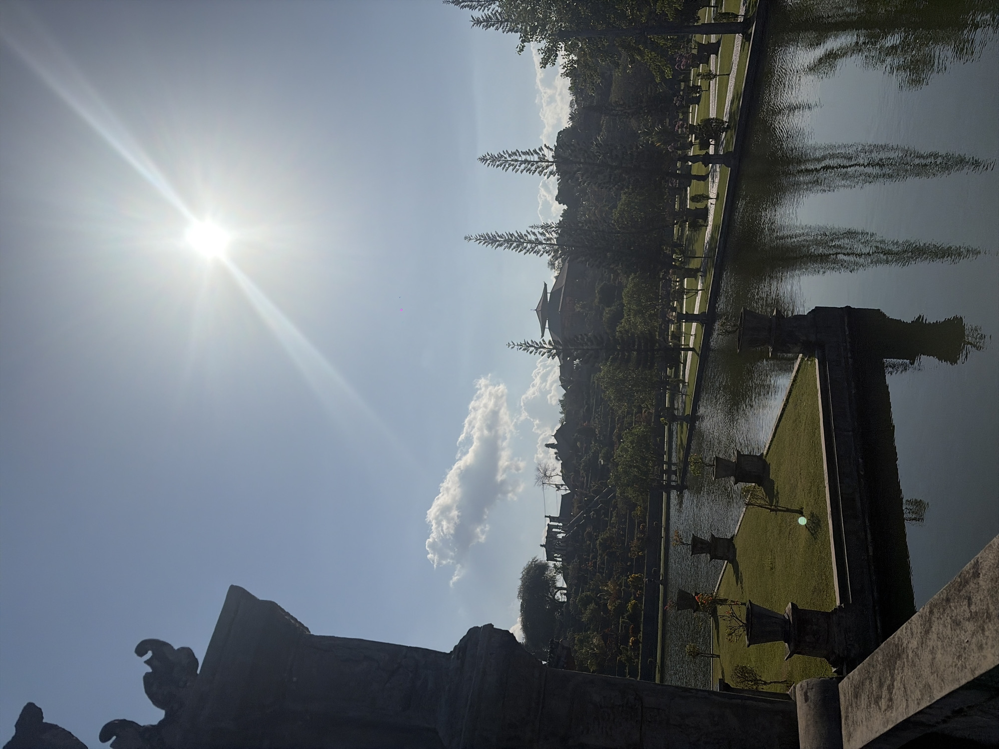 This is a vertical, medium-shot photograph of the Tirta Gangga Water Palace in Karangasem, Indonesia, taken on a bright, sunny day. The foreground is dominated by a stone railing and a portion of an ornate stone structure on the left, possibly part of a building or monument. Beyond this, a serene body of water reflects the sky and surrounding greenery.
The water is bordered by manicured green lawns and pathways. Several large, dark stone pots, some containing small trees or bushes, are strategically placed on raised platforms along the water's edge. In the mid-ground, the landscape rises, featuring terraced gardens with more trees and architectural elements, including what appears to be a multi-tiered building or pavilion with a dark roof.
The background is a lush green hillside dotted with trees and more structures, all bathed in the bright sunlight. Tall, slender coniferous trees are prominent in the mid-ground and background. The sky is a clear blue, with a few scattered white clouds and the sun shining intensely, creating a lens flare that emanates rays of light across the upper portion of the image. The overall scene evokes a sense of tranquility and grandeur, typical of a royal water garden. No people are visible in the image.