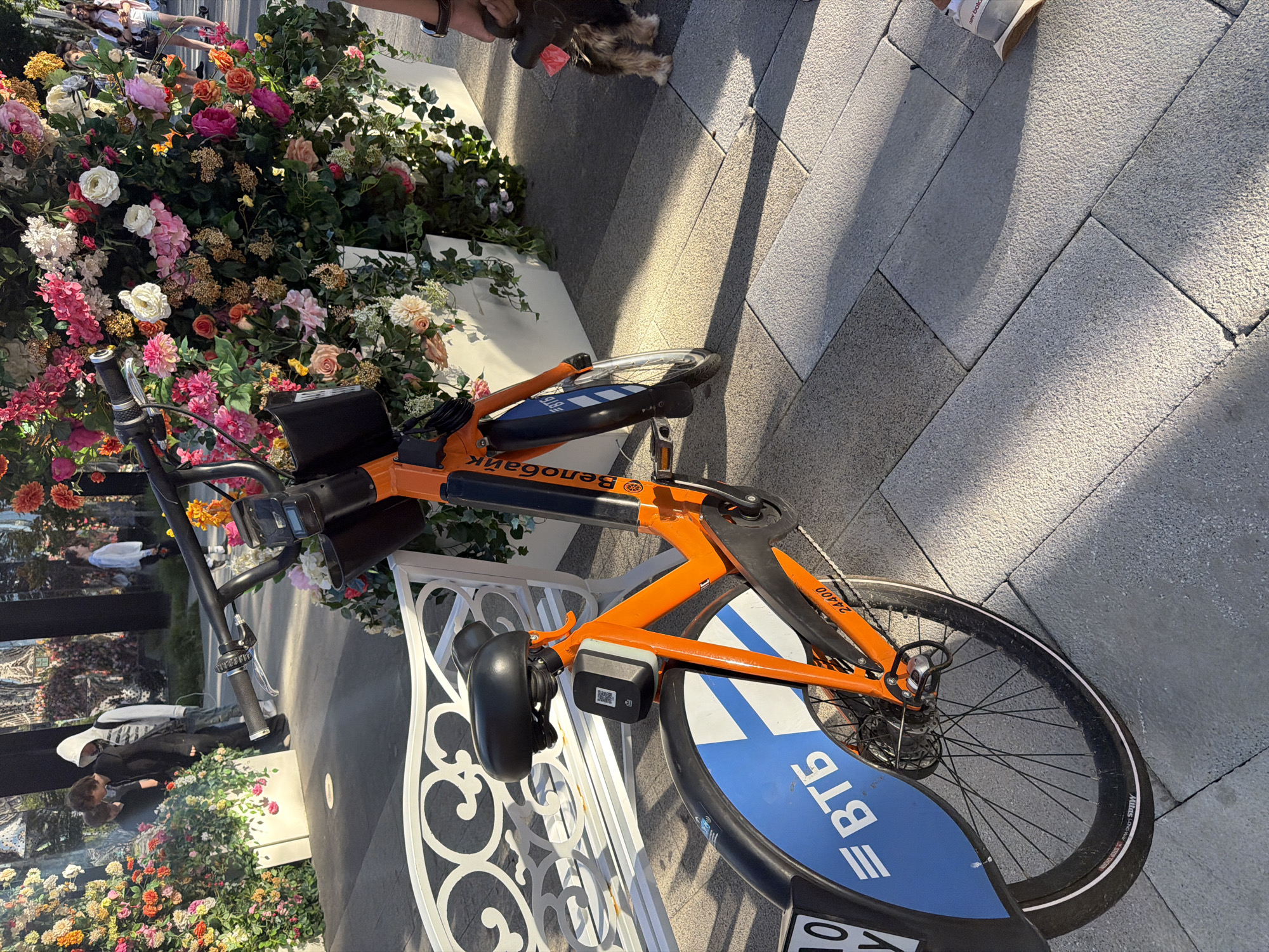 The image shows an orange bike with "Велобайк" written on its frame. The bike has a black seat and a QR code attached to its front. The bike is parked on a sidewalk made of gray paving stones. In the background, there is a white bench and a large floral arrangement with pink, orange, and white flowers. Several people are visible in the background, some of whom are looking at the flowers. A small dog on a leash is also present in the scene, being held by a person whose hand is visible. The lighting suggests it is daytime, with shadows cast by the bike and floral arrangement.