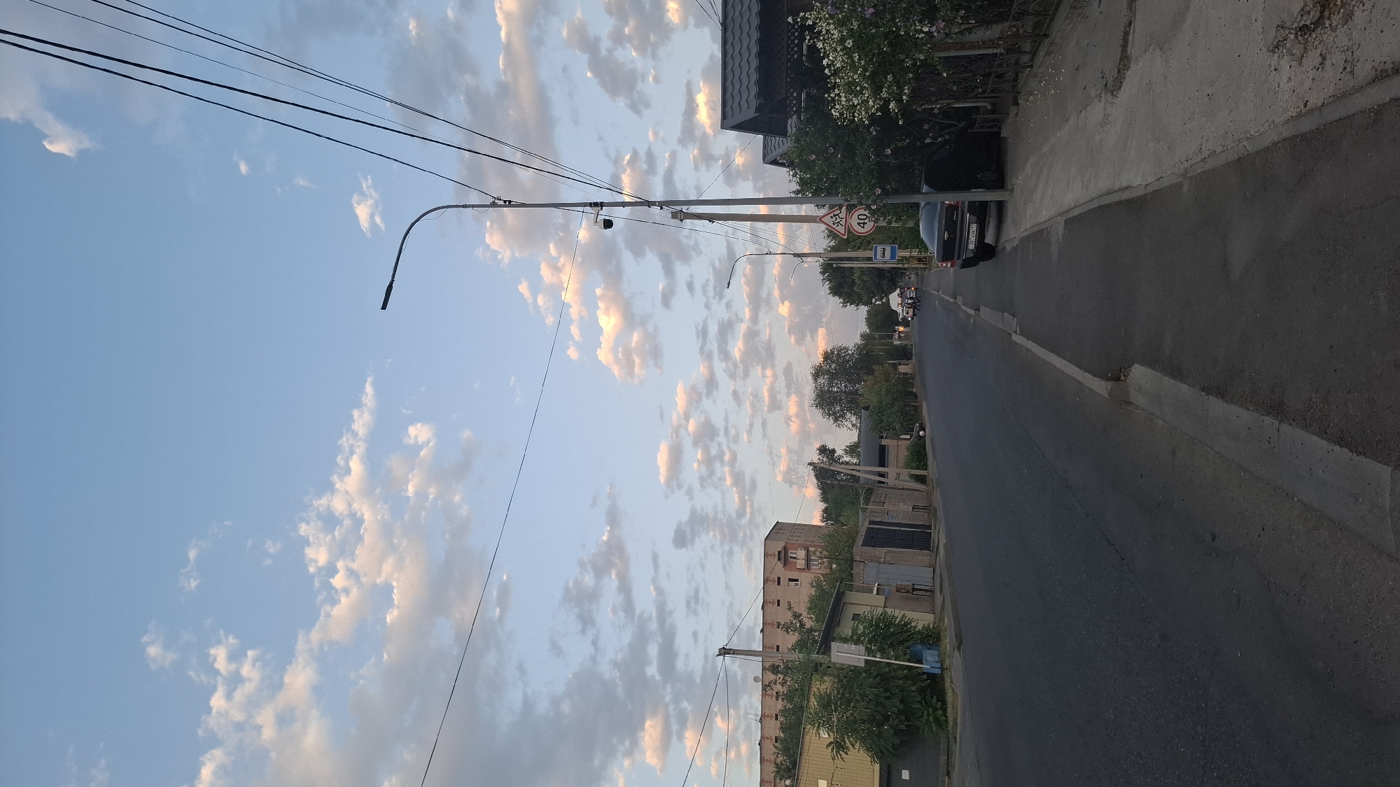 The image depicts a street scene in the city of Shymkent, Kazakhstan, under a partially cloudy sky. The perspective is from the sidewalk, looking down a paved road that curves slightly to the left and recedes into the distance. On the left side of the road, there are several low-rise buildings, including a yellow structure with a darker roof and a multi-story building with a patterned facade. Trees and bushes are visible between the buildings. On the right side of the road, a sidewalk runs parallel, with a black car parked alongside it. Further down the street, a sign indicating a speed limit of 40 is visible on a pole, along with a bus stop sign and a security camera. Power lines are strung across the upper portion of the image. In the distance, more trees and buildings are present, and several motorcycles are visible on the road. The overall impression is of a residential or mixed-use street during daylight.